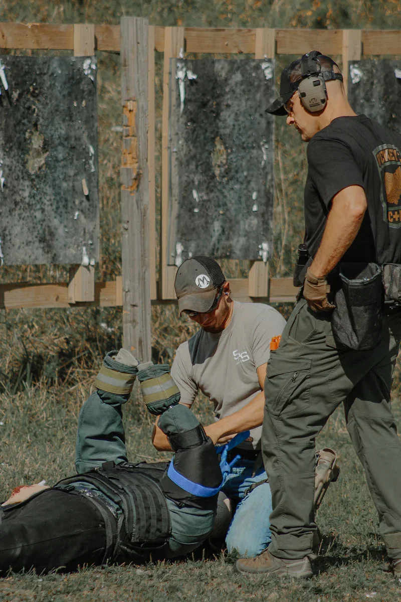 Two men assisting a person lying on the ground during a training exercise outdoors, with wooden targets in the background.