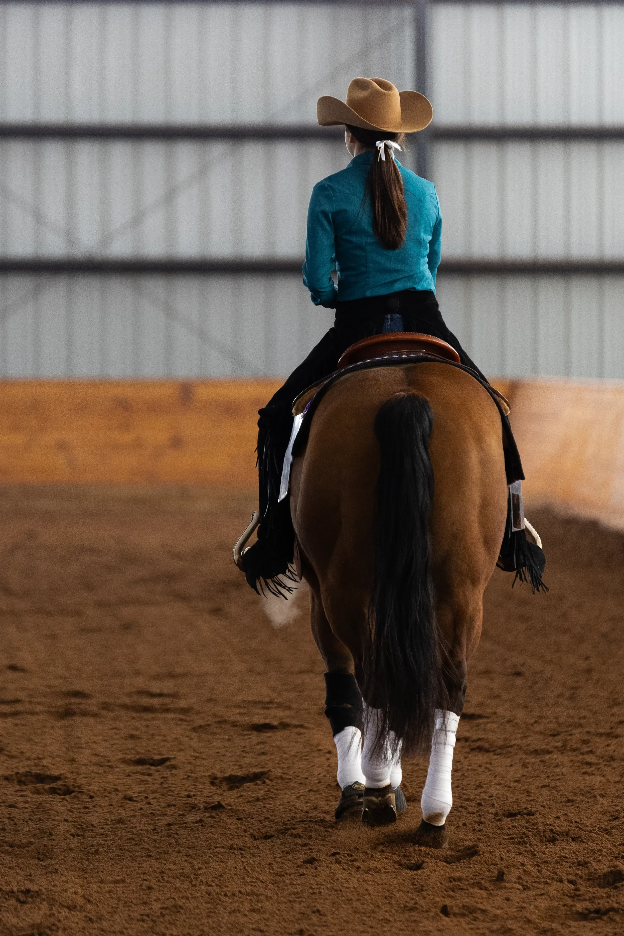 A woman riding a horse in an indoor arena, viewed from the back, wearing a cowboy hat, blue shirt, and black chaps.