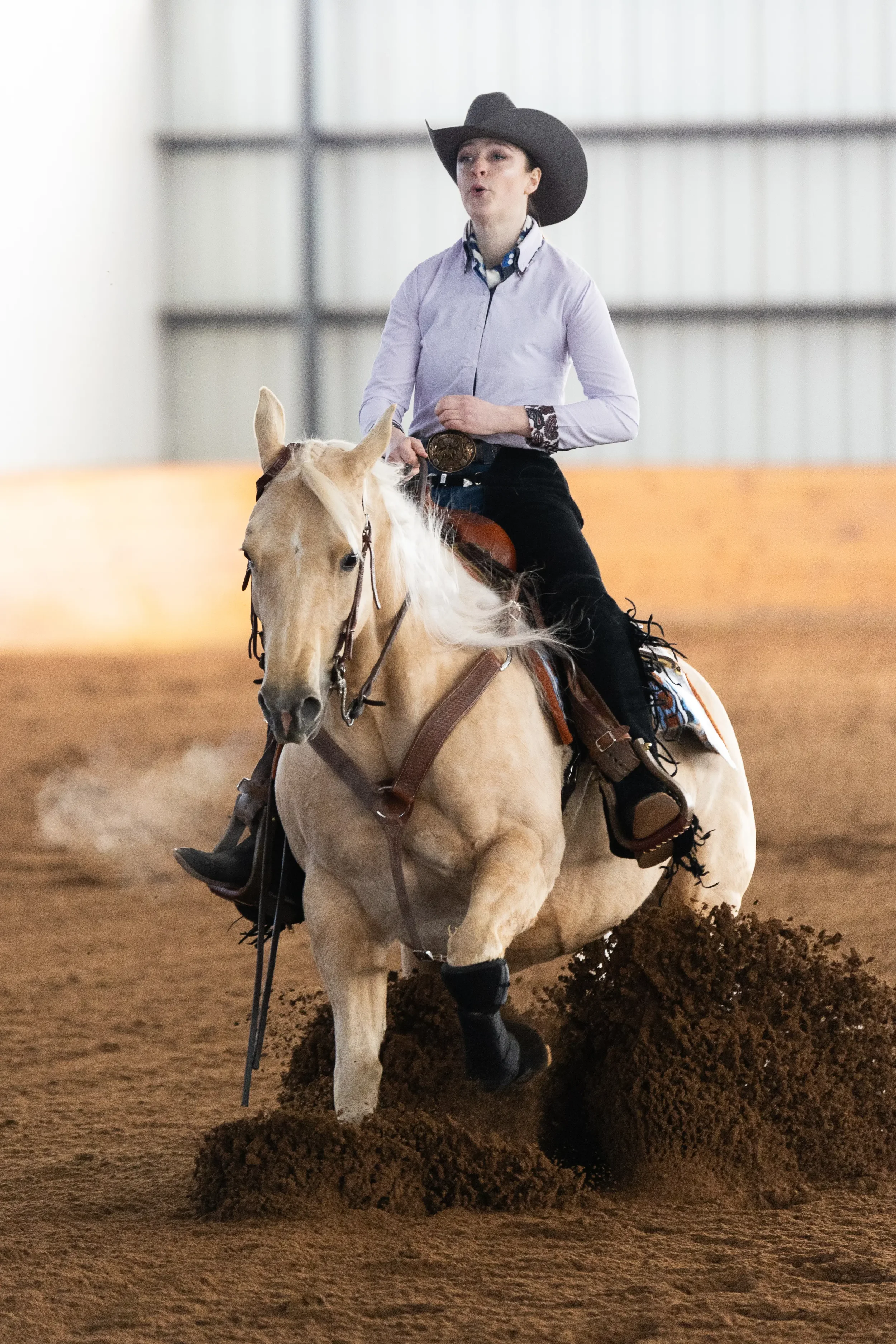 A woman wearing a black cowboy hat, a light purple shirt, and black pants riding a cream-colored horse in an indoor arena. The horse is kicking up dirt as it runs.