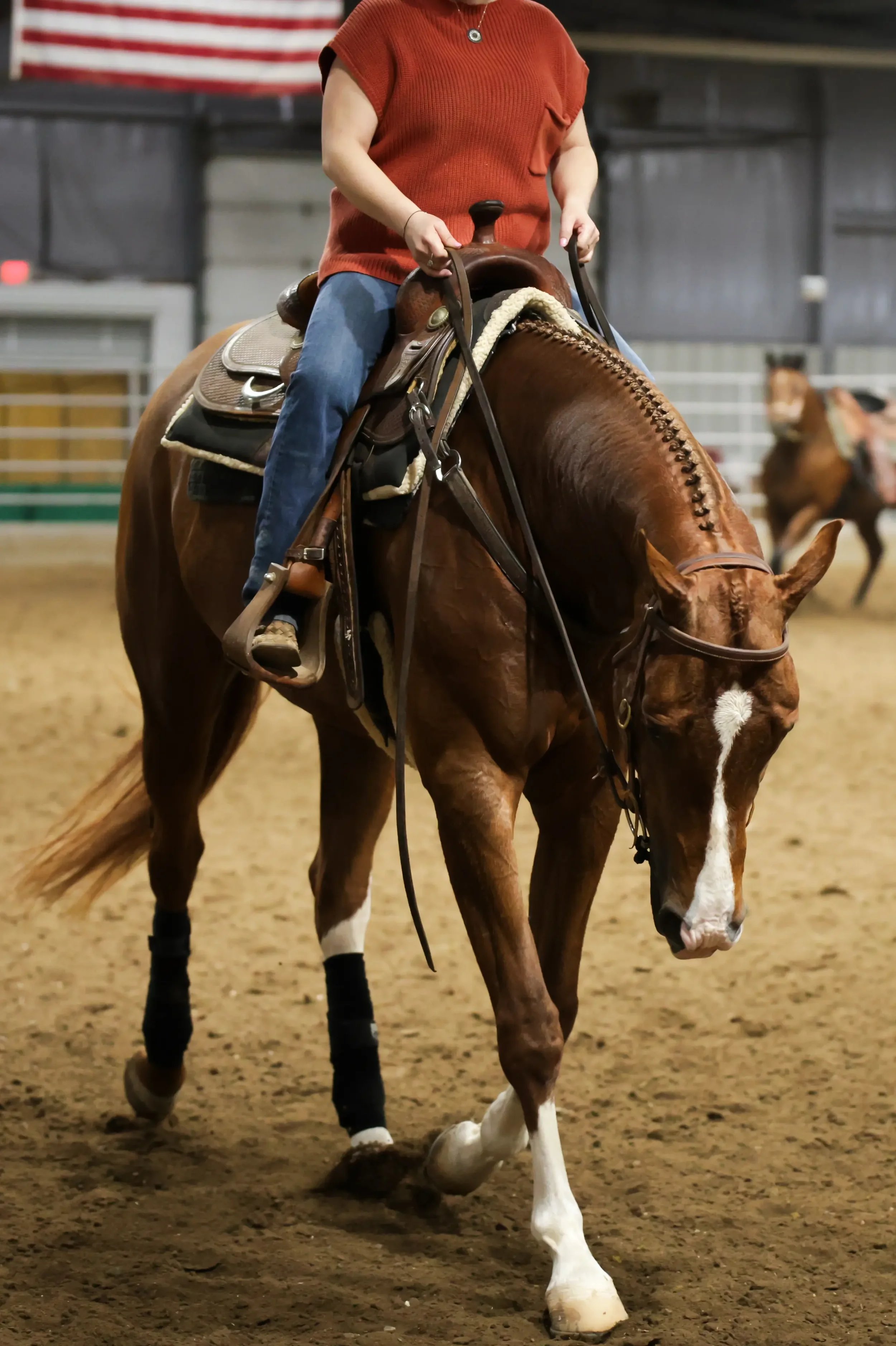 Person riding a brown horse with a white blaze, inside an indoor riding arena.