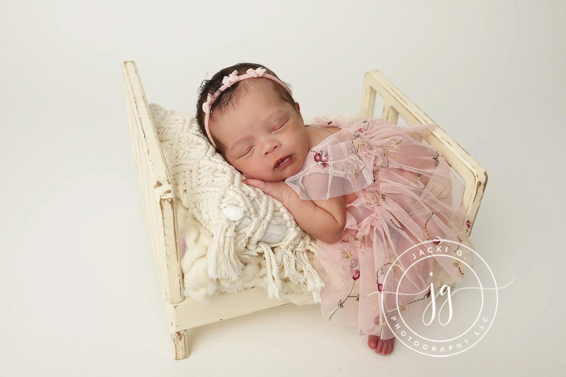 A sleeping infant in a pink dress with floral embroidery, lying on a white wooden bed with a knitted blanket and cream-colored pillows. The baby has a pink headband with small bows.