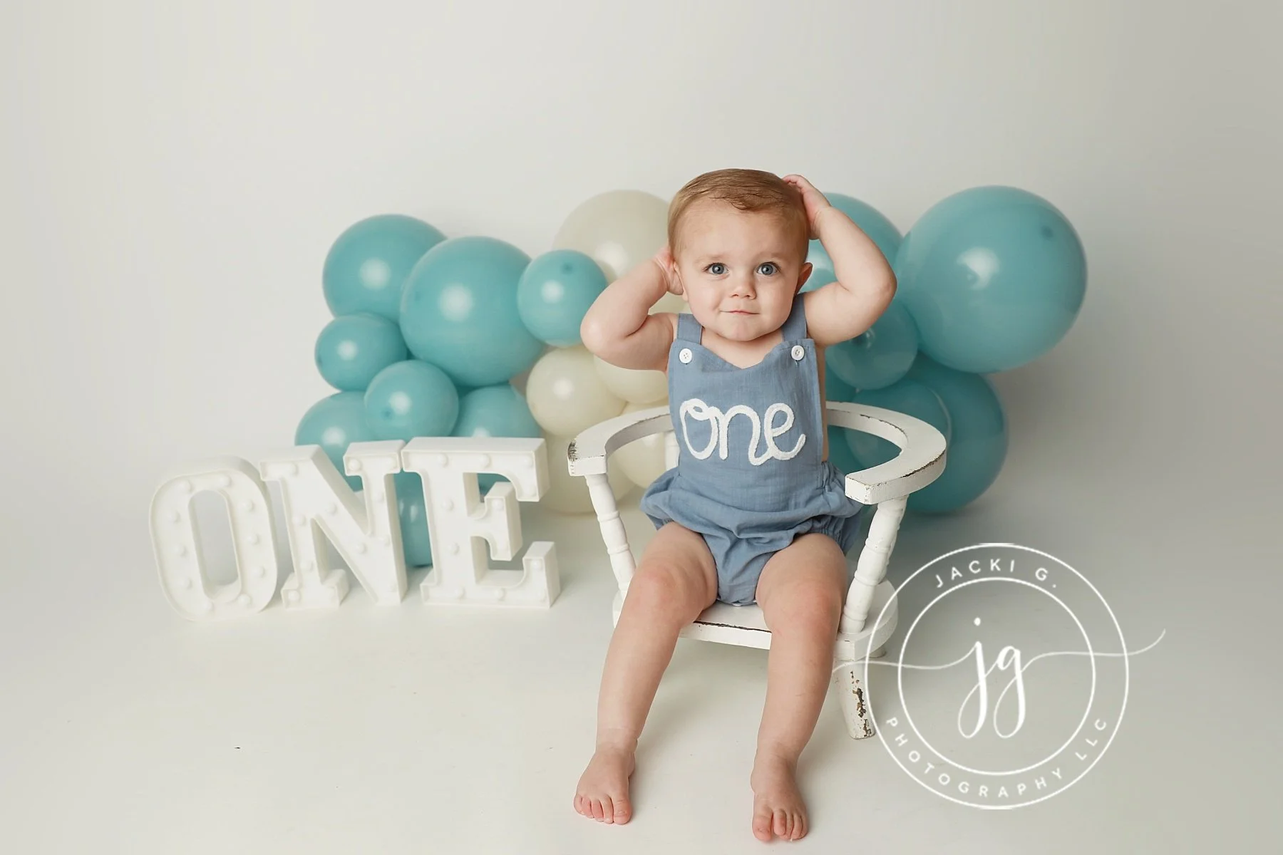 A baby with blue eyes and light hair sitting on a small white wooden chair, with one hand on their head and the other behind their ear, wearing a blue romper with the word 'one' on it. Behind the baby, there is a display of blue and white balloons, large illuminated white letter spellings of 'ONE,' and a circular watermark from a photography studio in the bottom right corner.