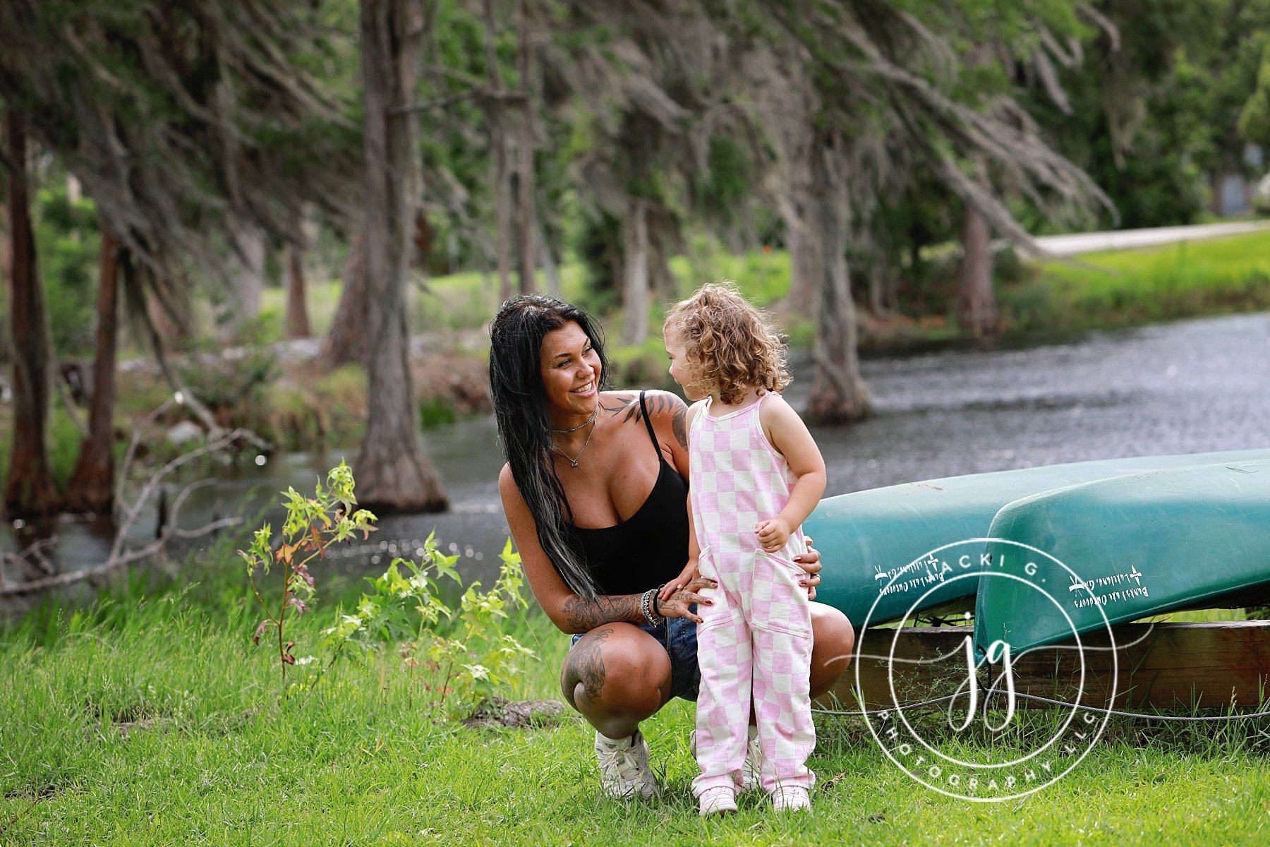 A woman with long dark hair and tattoos kneels next to a young girl with curly blonde hair, both smiling and looking at each other near a green canoe by a river, surrounded by trees and green grass.