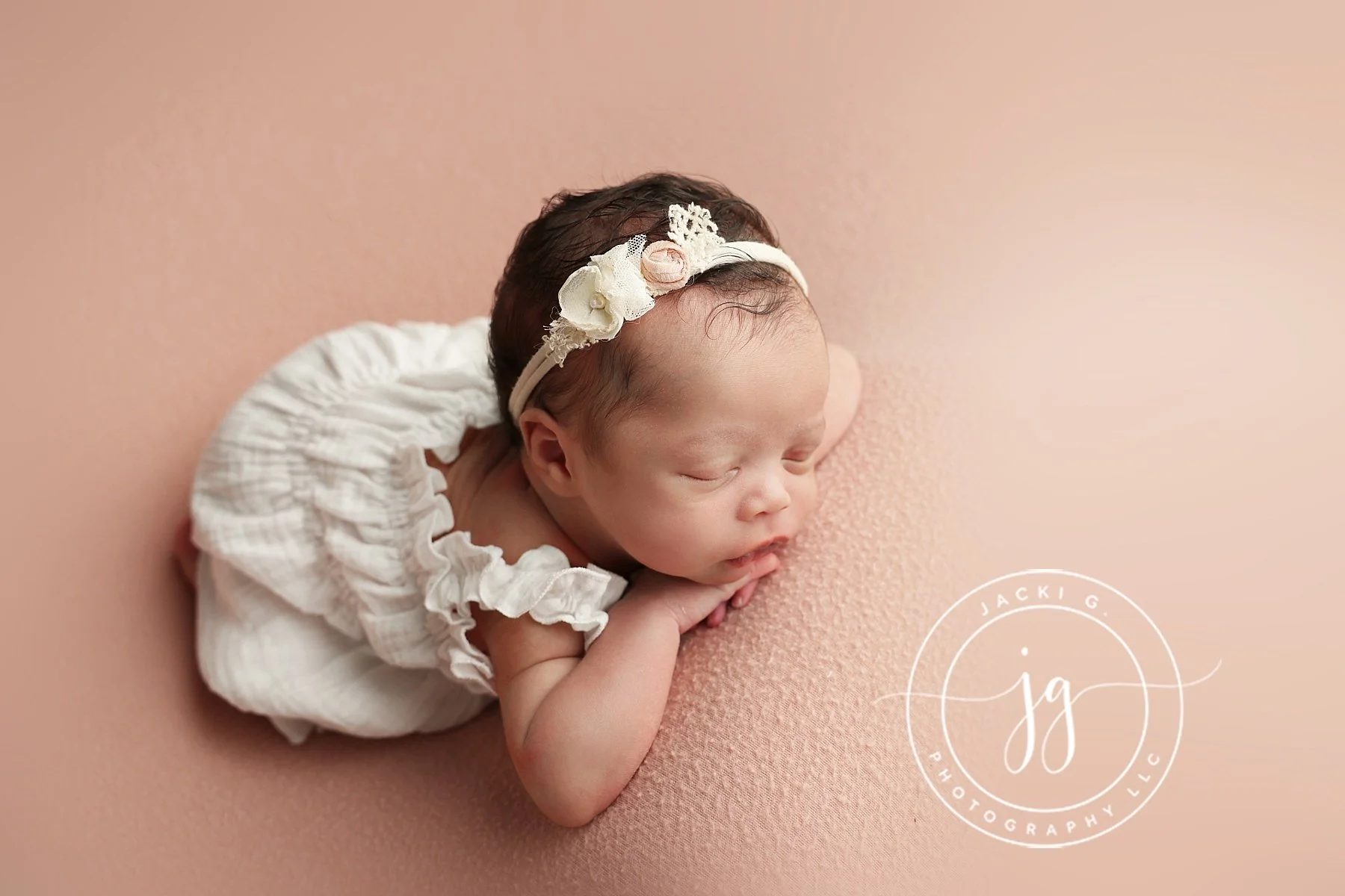Newborn baby with dark hair, sleeping on a soft pink surface, wearing a cream-colored headband with flowers, dressed in a ruffled cream top, with hands folded under chin.