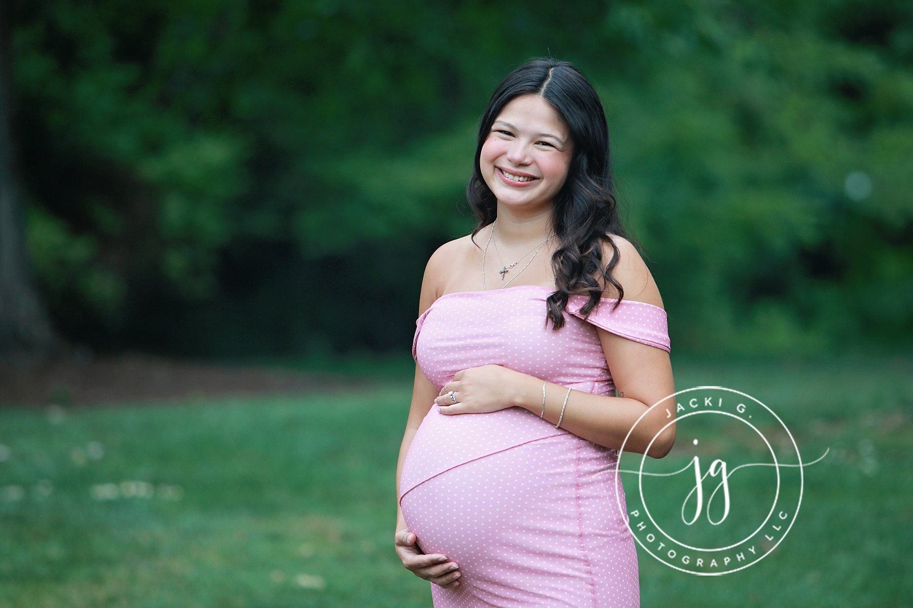 A pregnant woman with black hair, smiling, wearing a pink off-shoulder dress with white polka dots, standing outdoors on green grass and trees, with her hand on her belly.