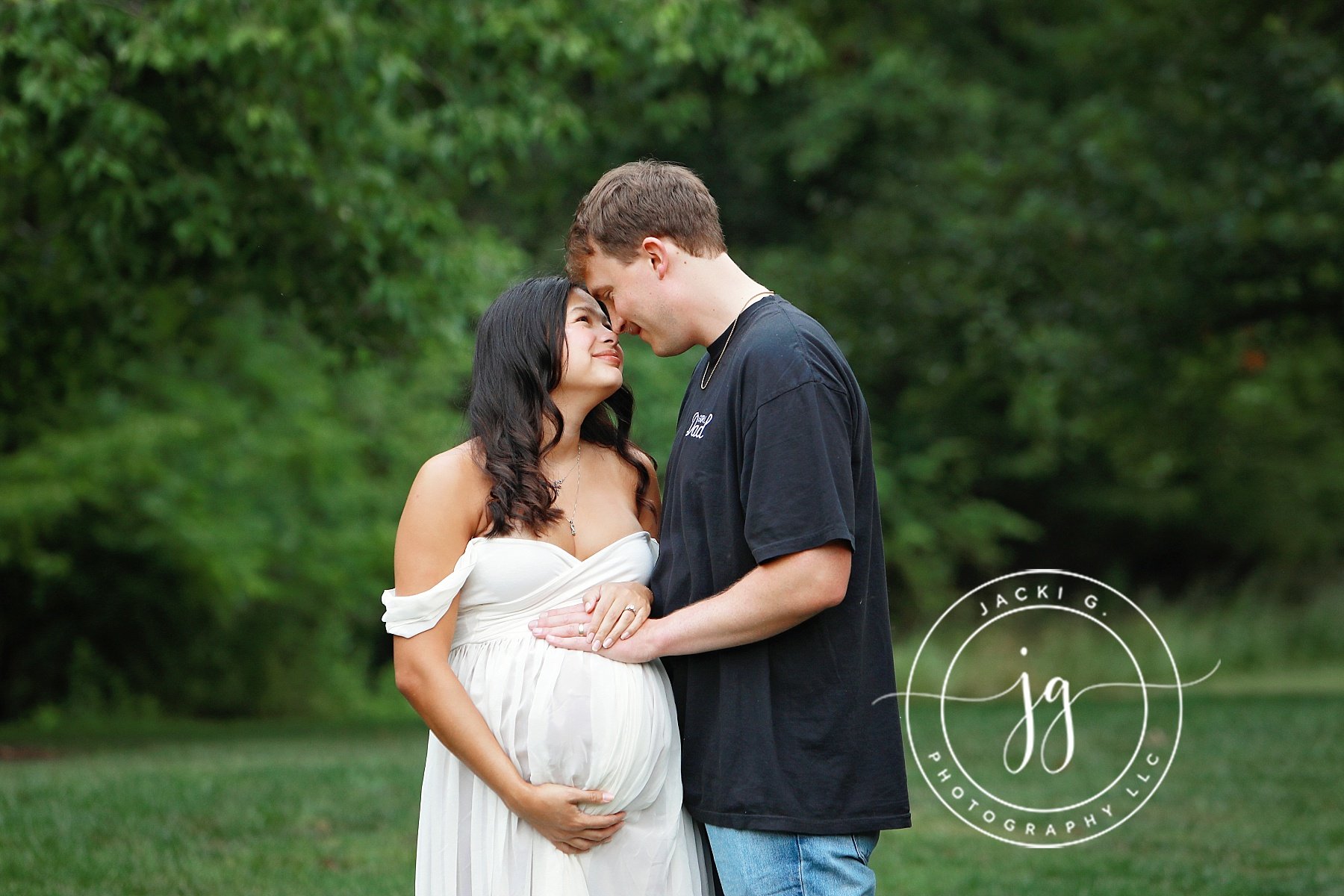 A pregnant woman in a white dress and a man with brown hair in a black t-shirt, standing close and touching foreheads in a green outdoor setting.
