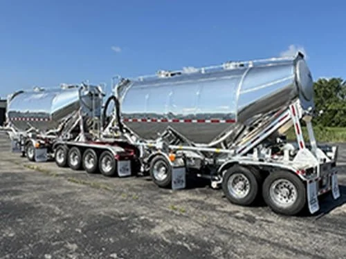 Two silver bulk liquid tank trailers connected together on a paved surface under a clear blue sky.