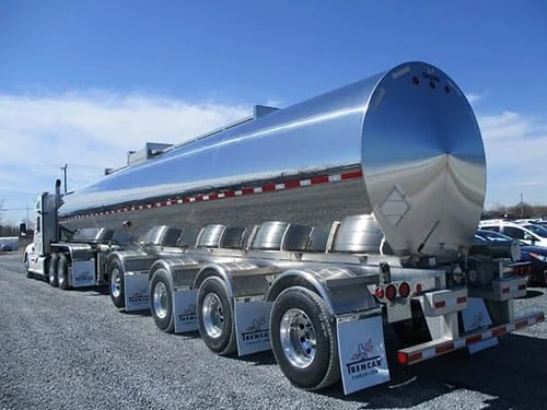 Silver tanker truck with multiple axles parked in a gravel lot under a clear blue sky.