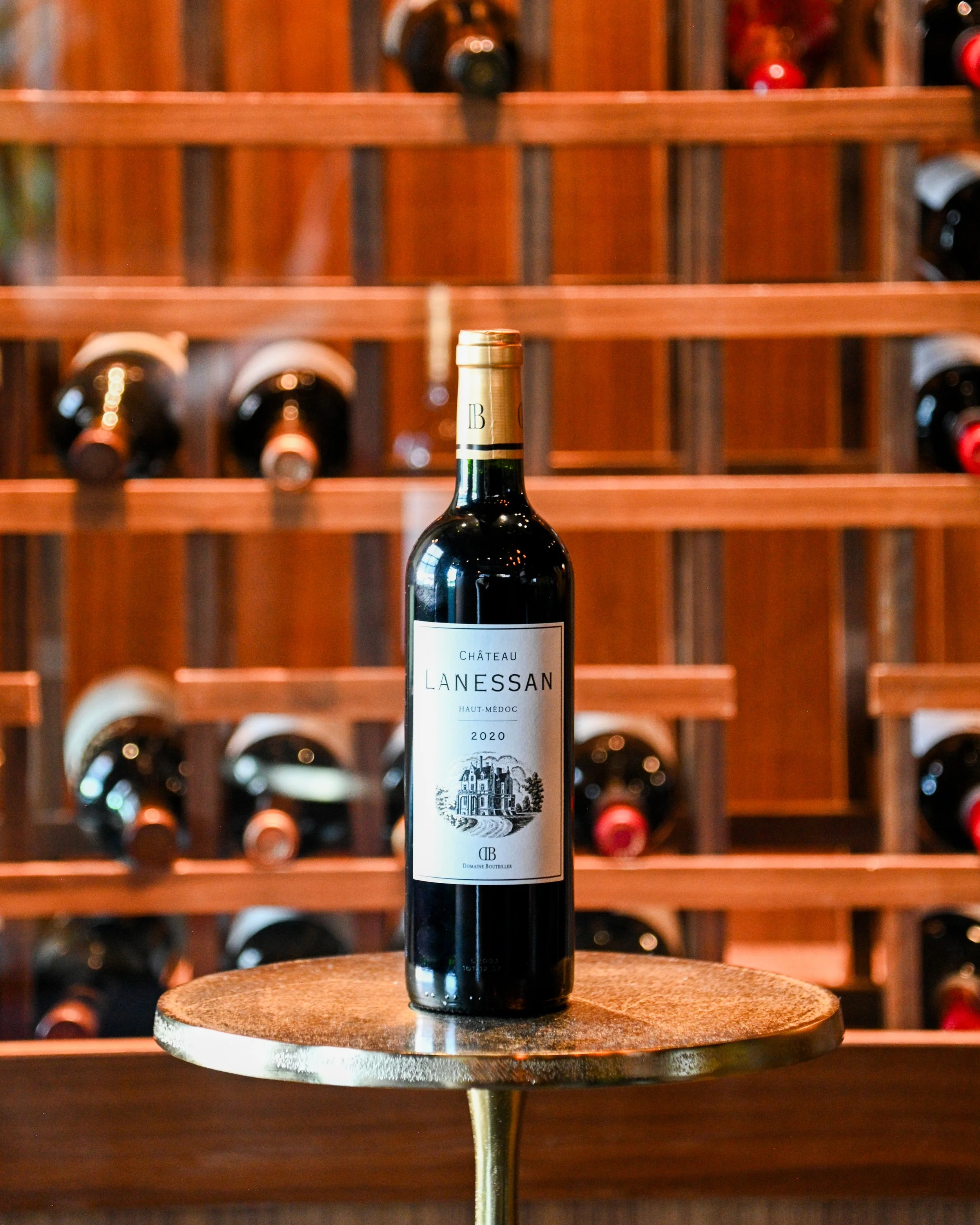 A bottle of Chateau Lanessan wine from 2020 standing on a small round wooden table in front of a wine rack filled with wine bottles in a wine cellar or store.