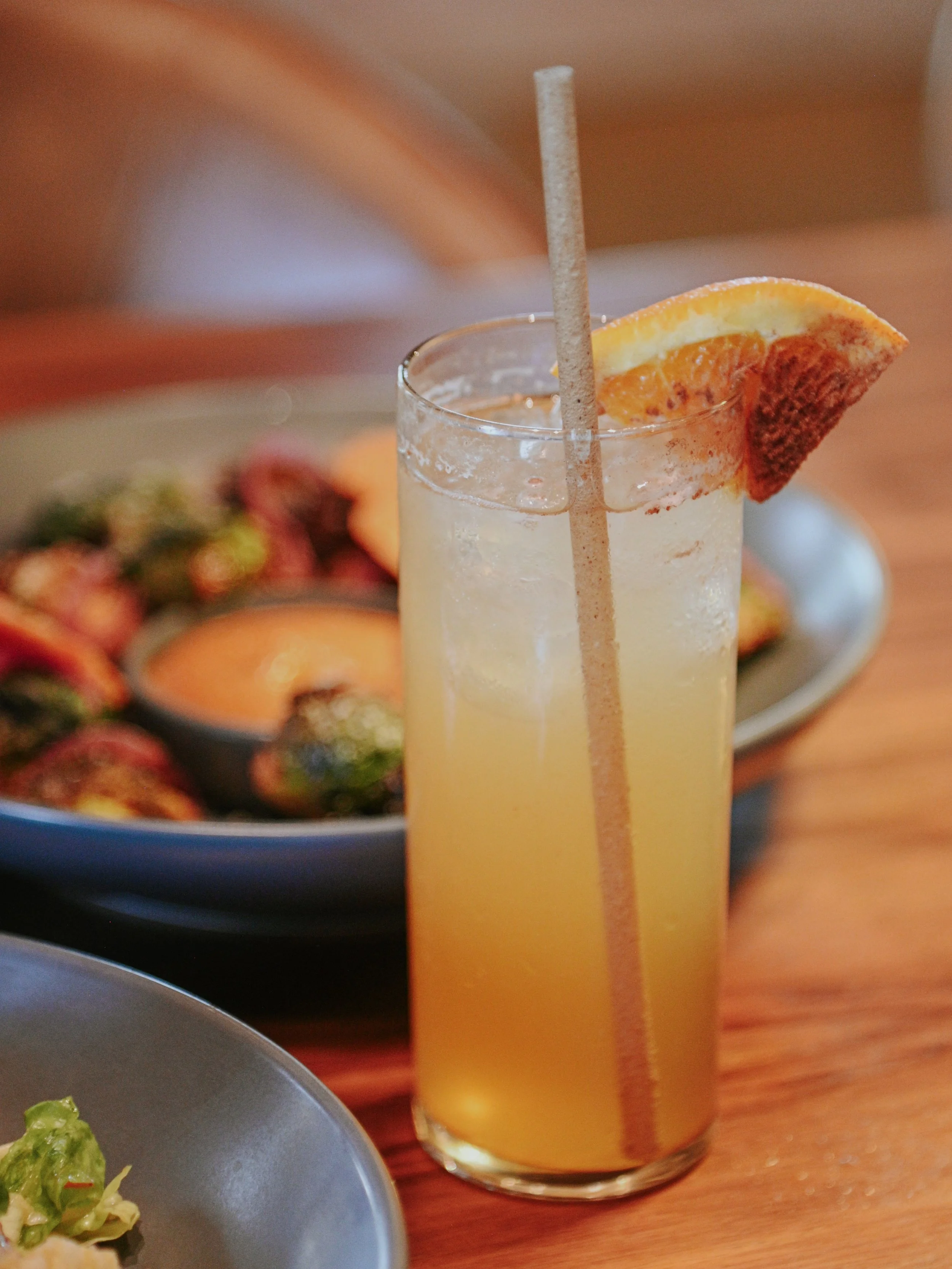 A tall glass of orange-colored drink garnished with a slice of orange and a straw, with food plates in the background.
