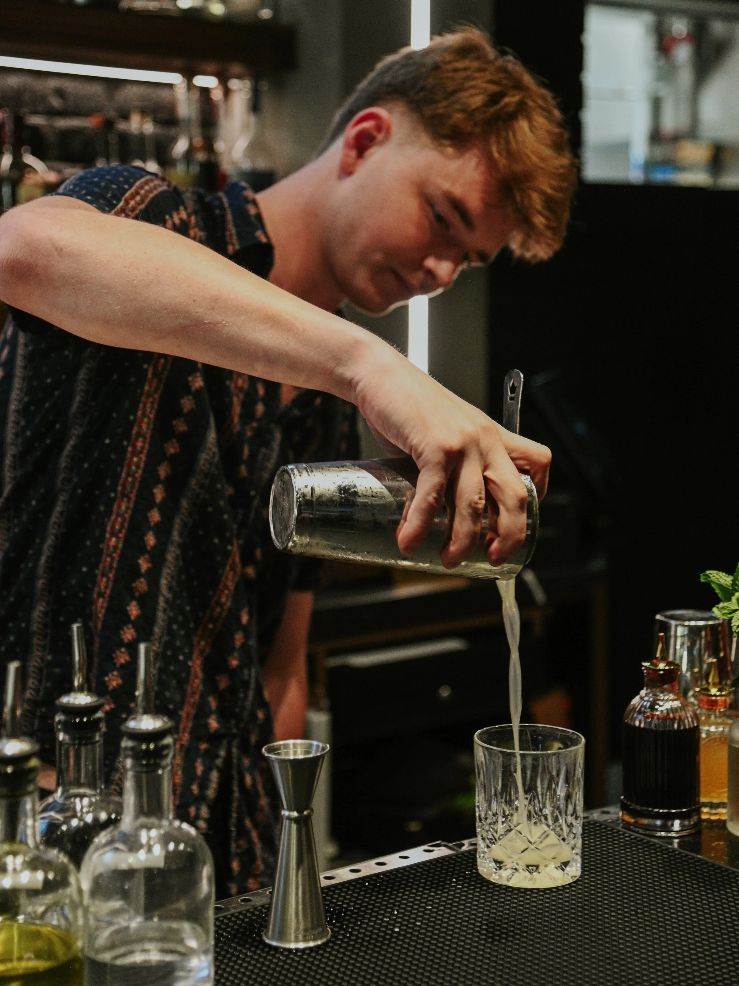 A bartender pouring a drink into a glass at a bar with bottles and bar tools on the counter.