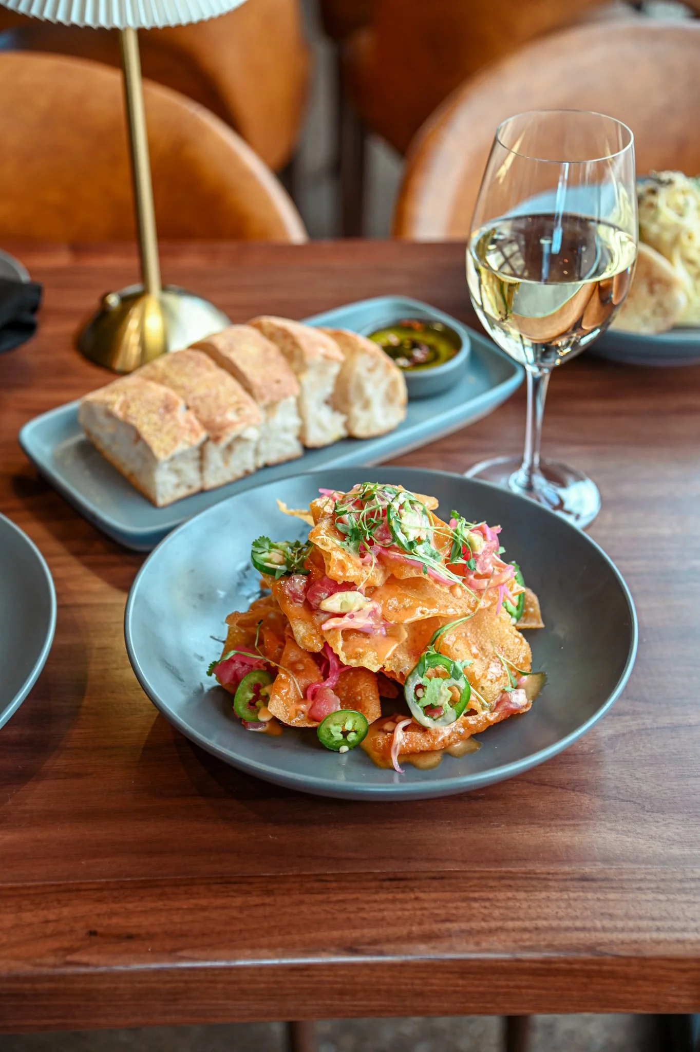Plate of nachos topped with sliced jalapeños, diced tomatoes, and microgreens, on a wooden table. Behind, a small loaf of bread and a glass of white wine.