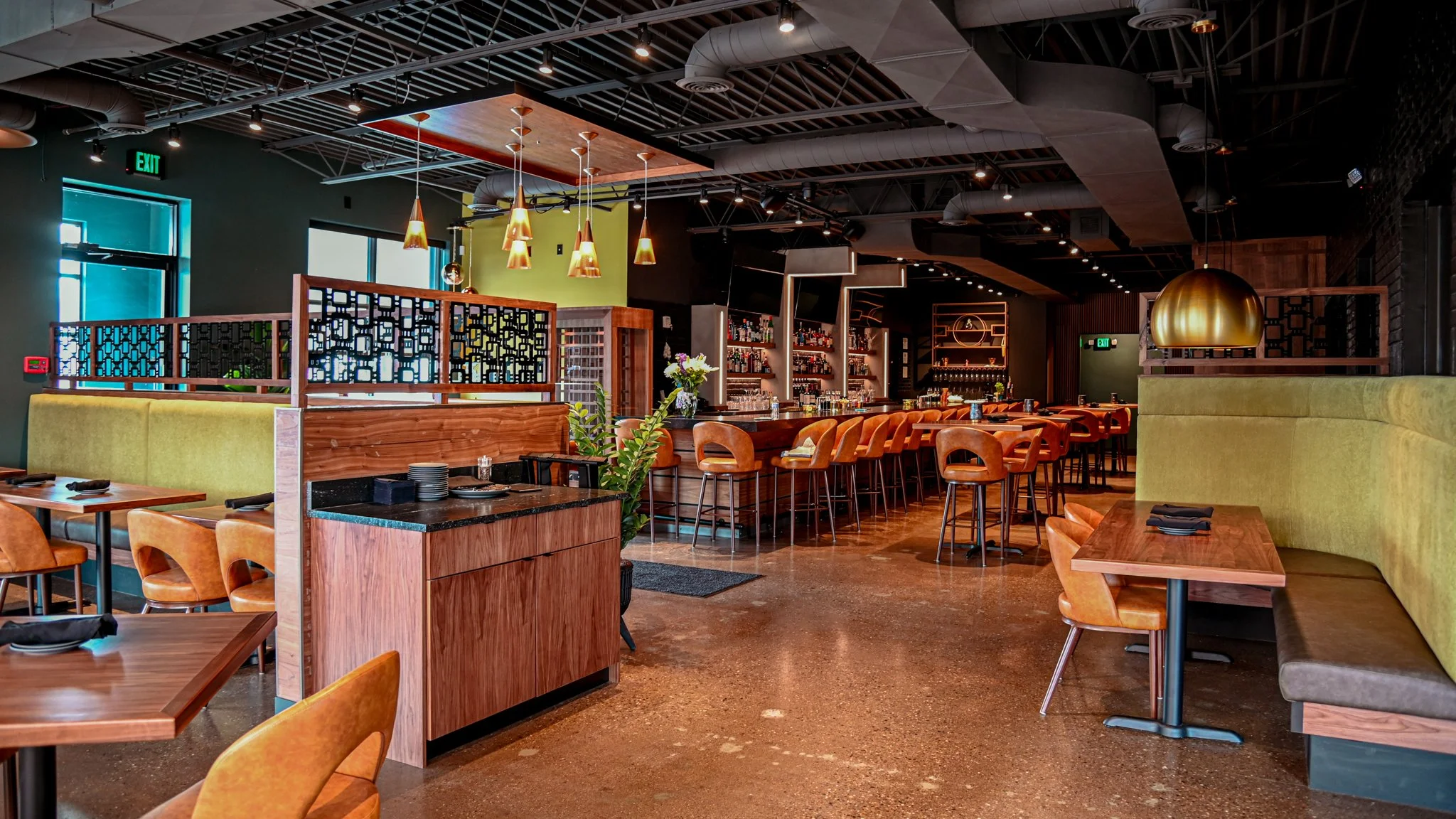 Interior of a modern restaurant or bar with wooden tables and leather chairs, bar counter with bar stools, shelves with bottles, green and black walls, and pendant lighting.