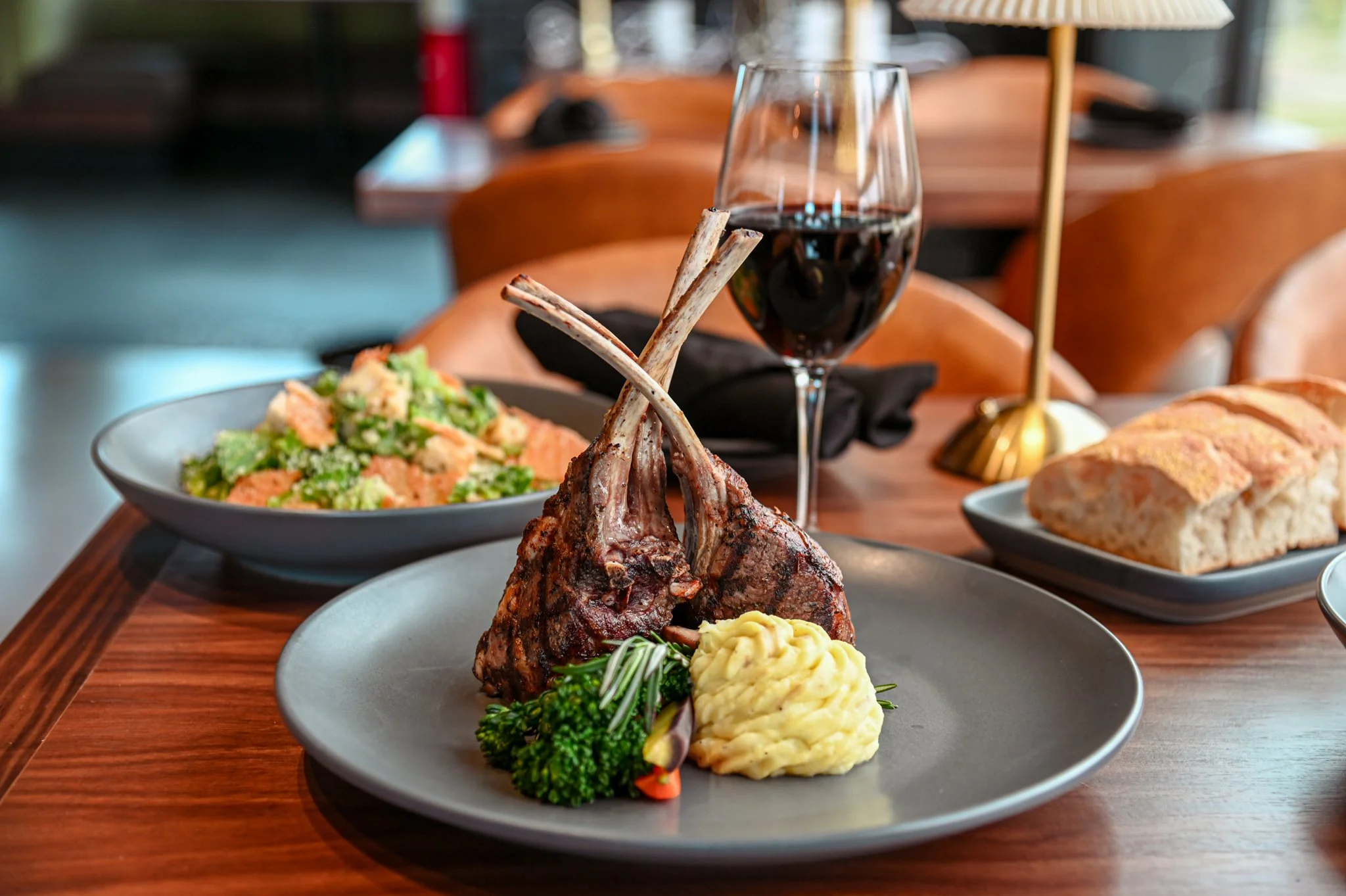 Plate with grilled lamb chops, mashed potatoes, and vegetables, alongside a glass of red wine and a bowl of salad on a wooden table.
