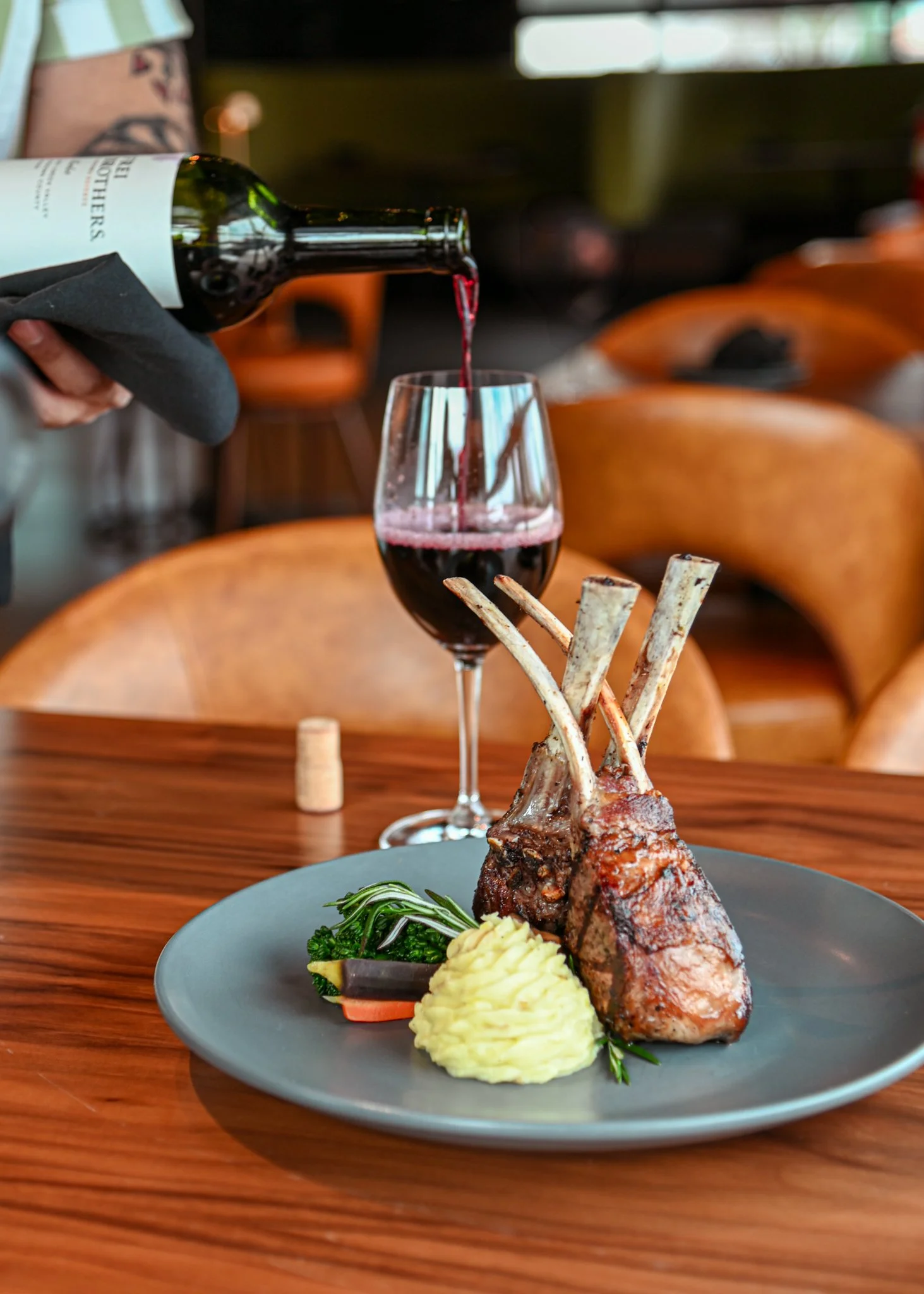 A restaurant meal with two grilled lamb chops, mashed potatoes, and vegetables on a gray plate, with red wine being poured from a bottle into a glass behind the plate.