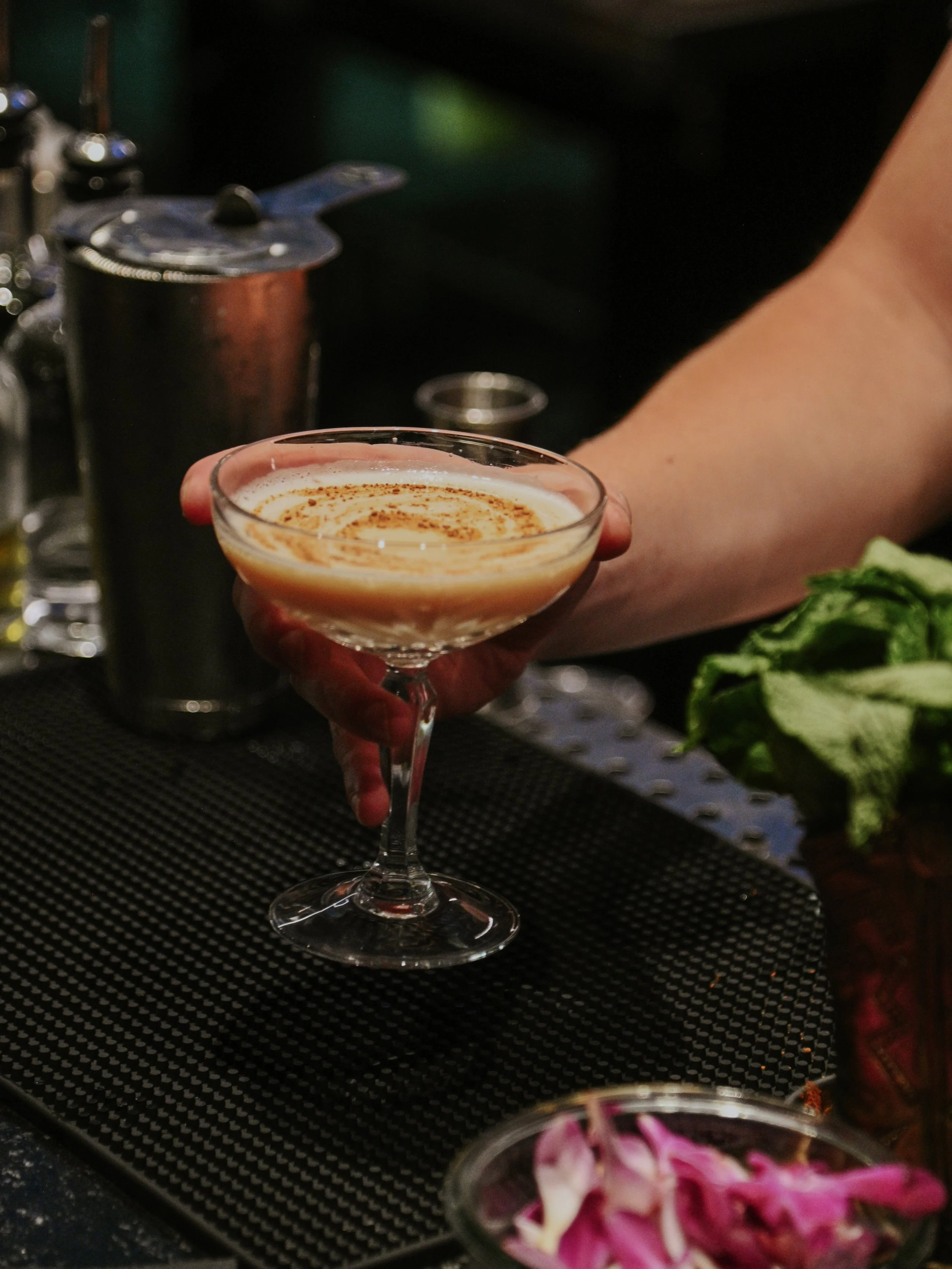 A bartender's hand holding a cocktail glass with a creamy, frothy drink garnished with a sprinkle of cinnamon, on a bar counter with various bar tools and a bowl of pink and white flower petals.