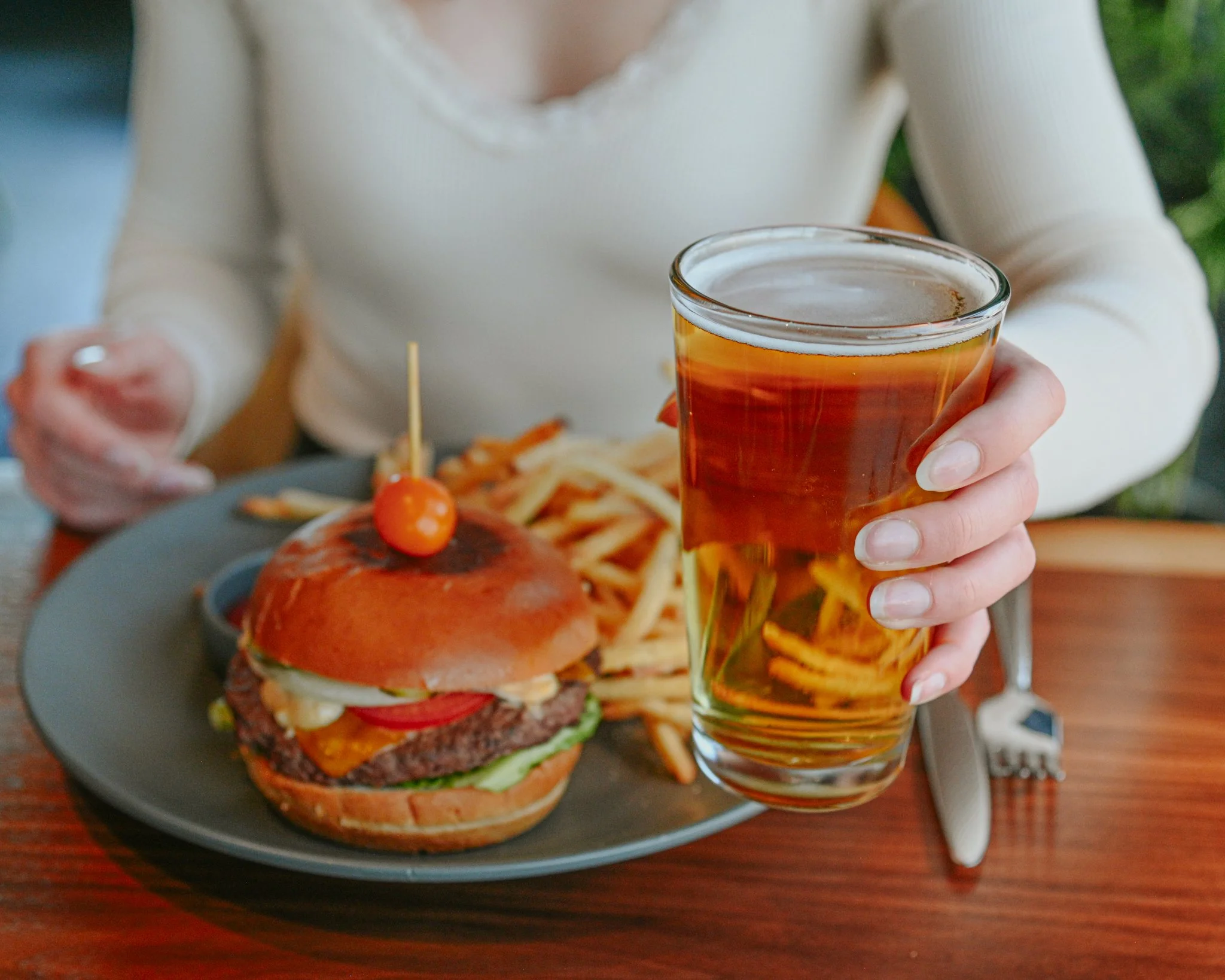A person holding a pint of beer with a plate of burger and fries on a wooden table.