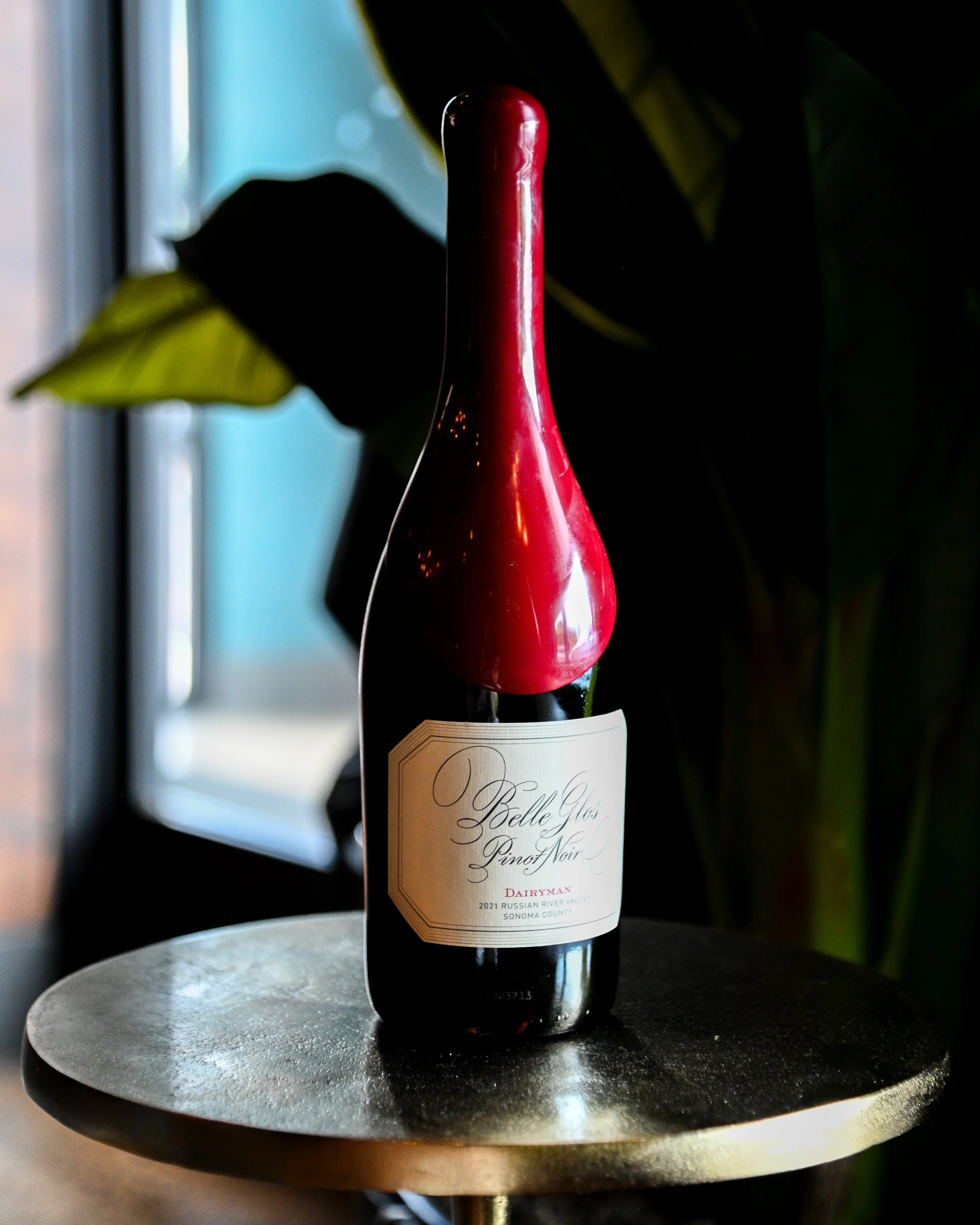 A wine bottle with a unique elongated, curved red wax seal on the top, placed on a dark round table in a dimly lit indoor setting with green plant leaves in the background.