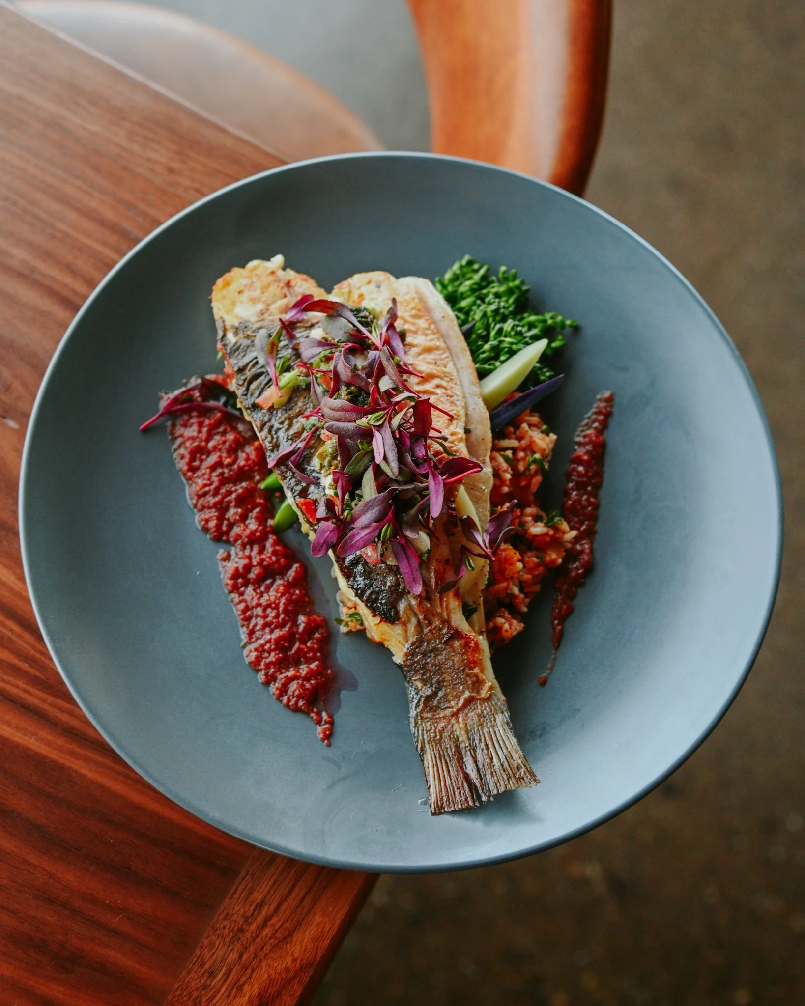 A plated dish with a whole grilled fish topped with purple microgreens, served with red sauce, green vegetables, and a grain side on a blue plate.