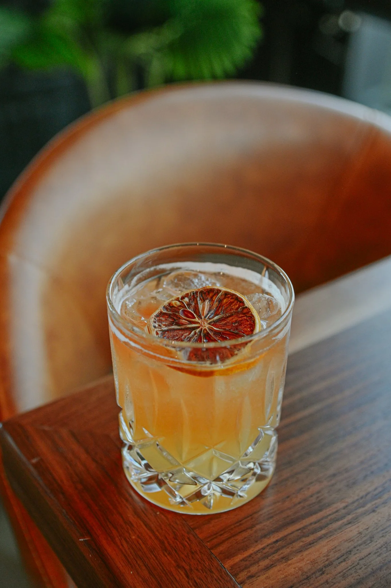 A glass of light brown beverage with ice and a slice of dried citrus fruit on top, on a wooden table.