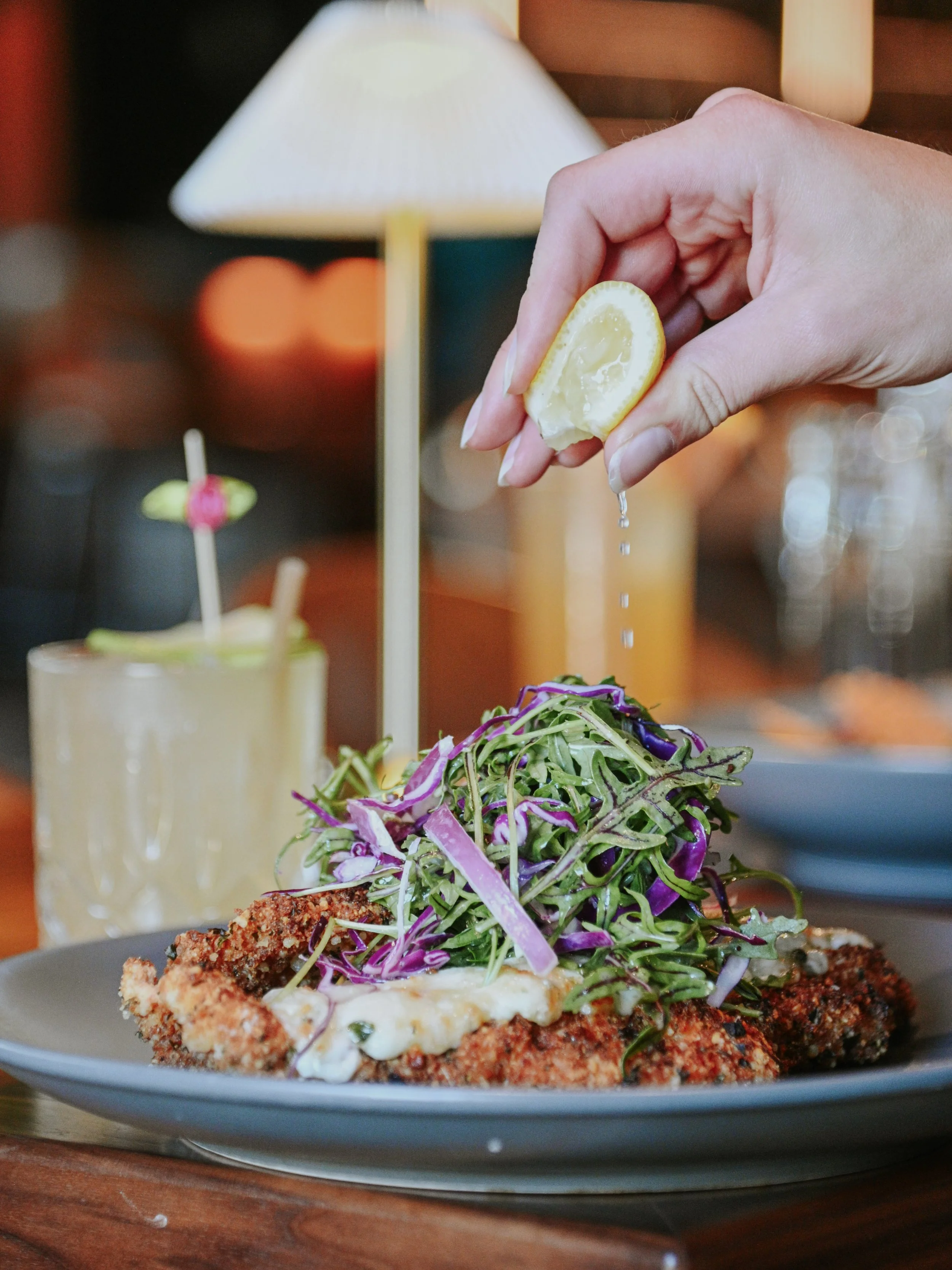 Hand squeezing lemon over a plate of fried chicken topped with a colorful salad. In the background, there are two beverages with garnishes and a lamp, in a cozy restaurant setting.