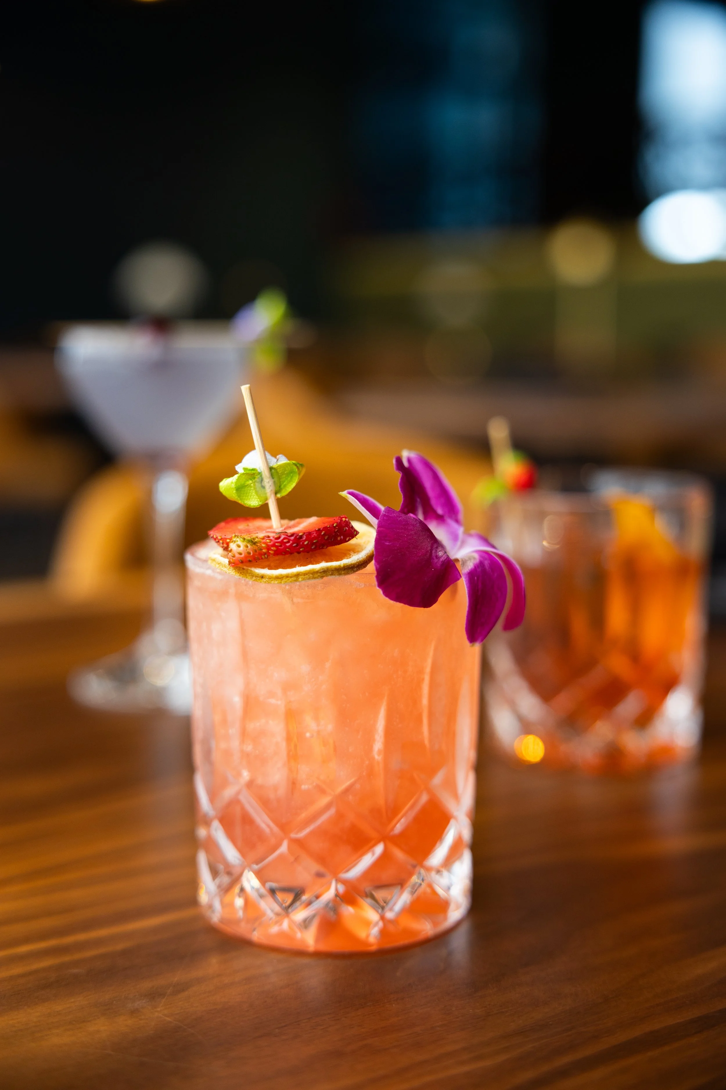 A pink cocktail garnished with a slice of strawberry, a slice of lemon, a green leaf, and a purple orchid, with a blurred background of other drinks on a wooden table.