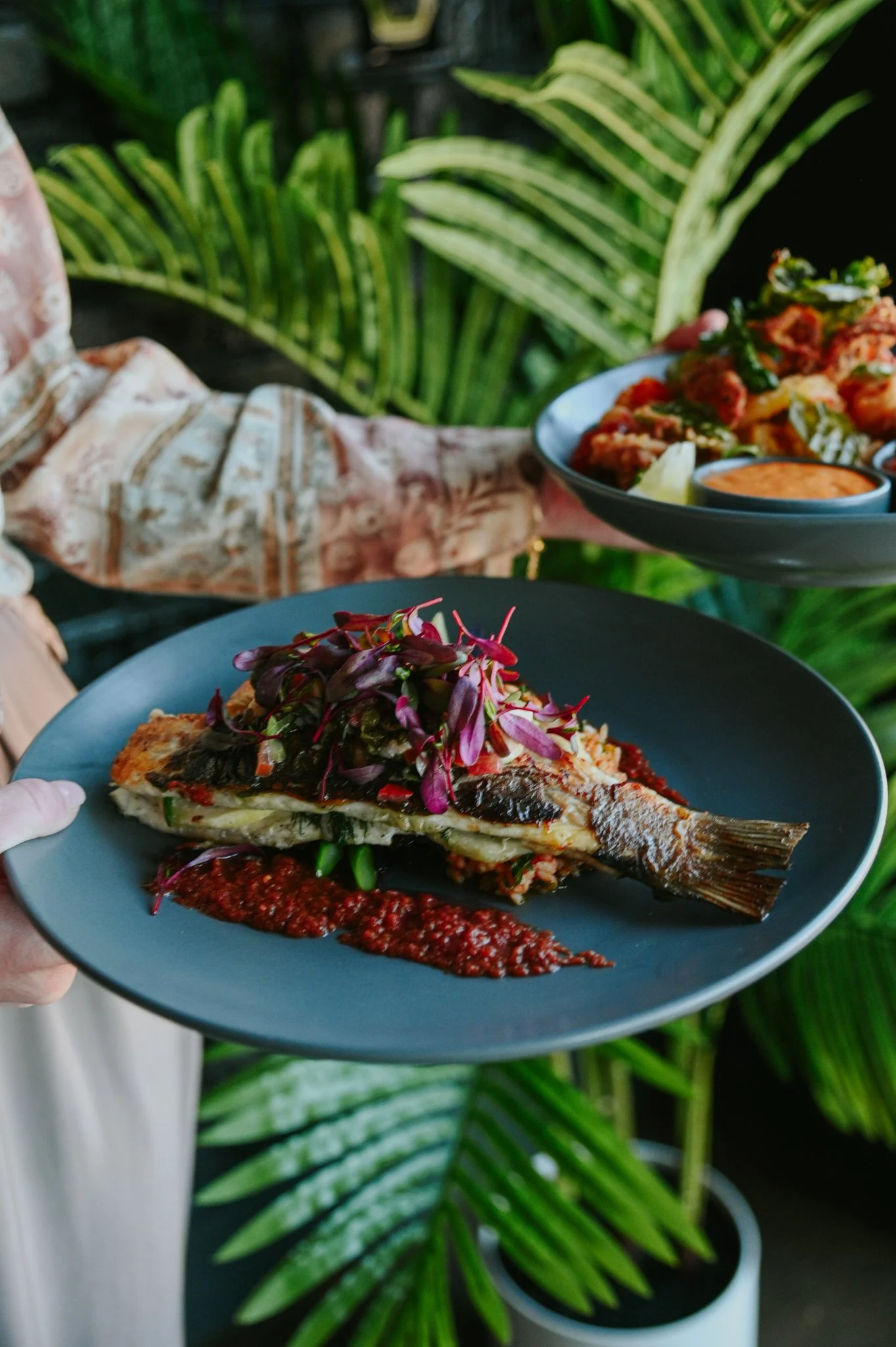 Person holding a black plate with a cooked fish fillet topped with vegetables and garnished with microgreens, accompanied by a red sauce on the side. In the background, a person is serving other dishes on a platter, with green tropical plants.
