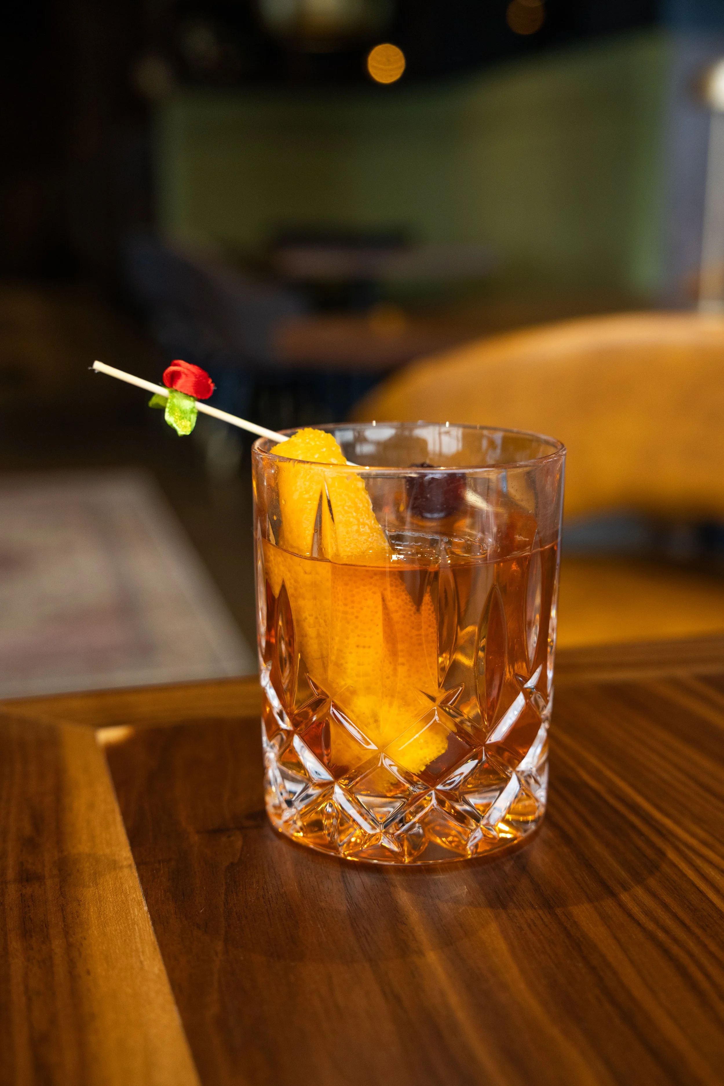A crystal glass filled with an amber-colored cocktail, garnished with orange slices and a cherry, on a wooden table in a cozy indoor setting.
