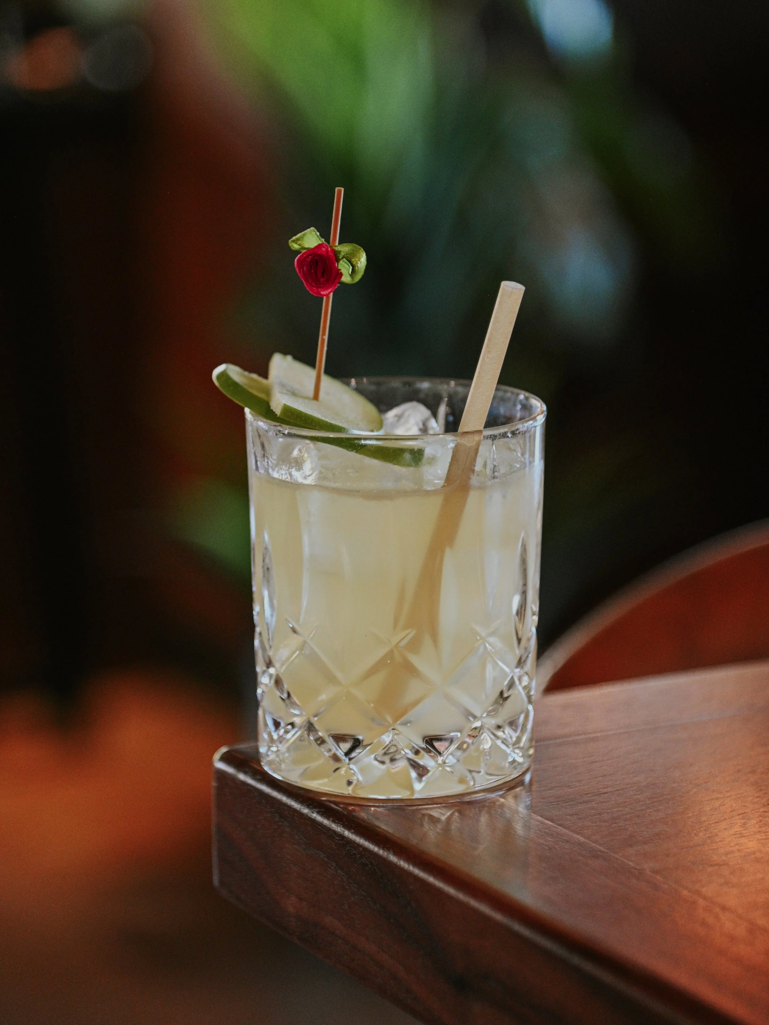 A cocktail in a faceted glass with lime wedges, a cherry, and a straw, placed on a wooden surface with a blurred green background.