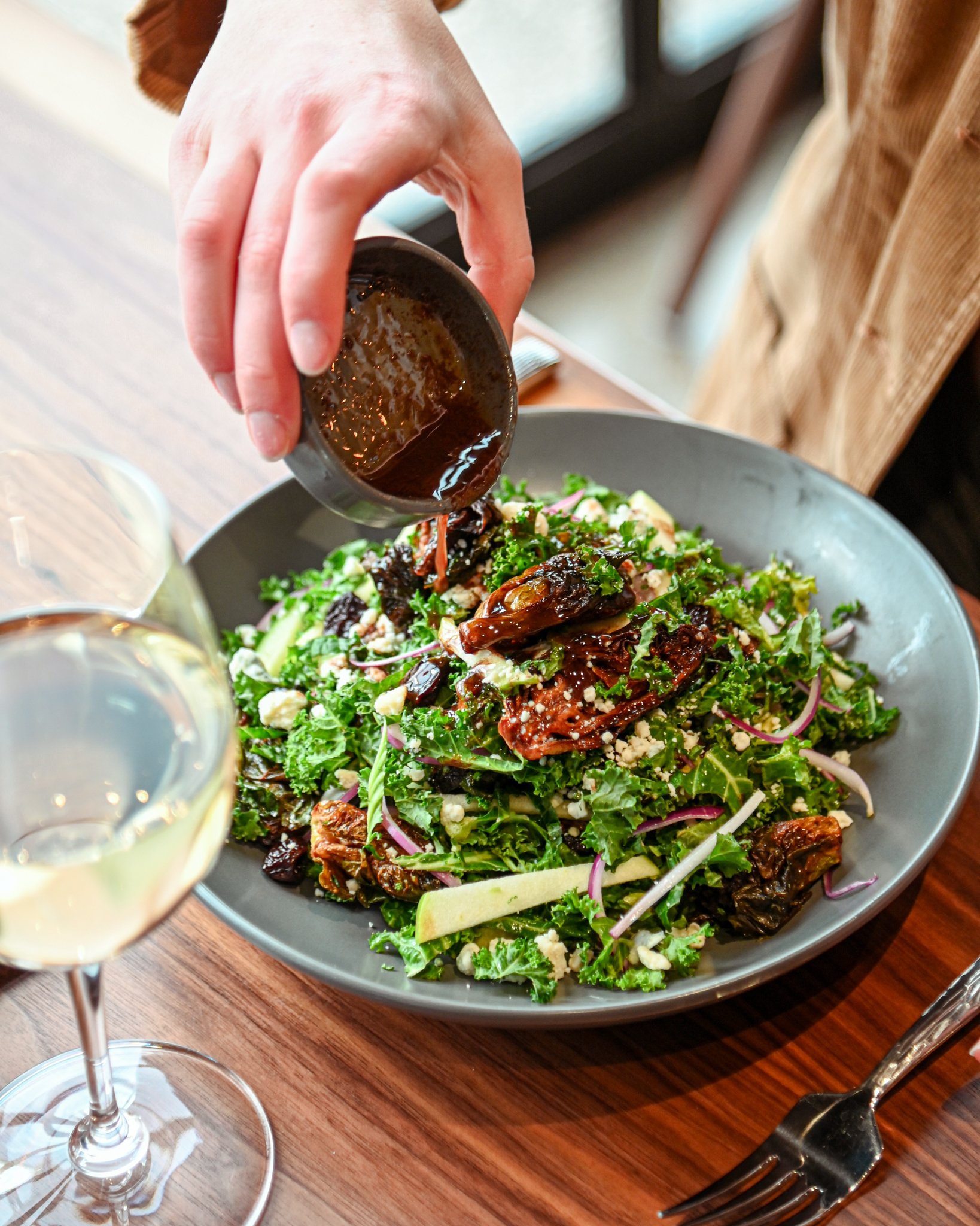 Person pouring dressing over a salad with greens, olives, cheese, and sun-dried tomatoes, with a glass of white wine on a wooden table.