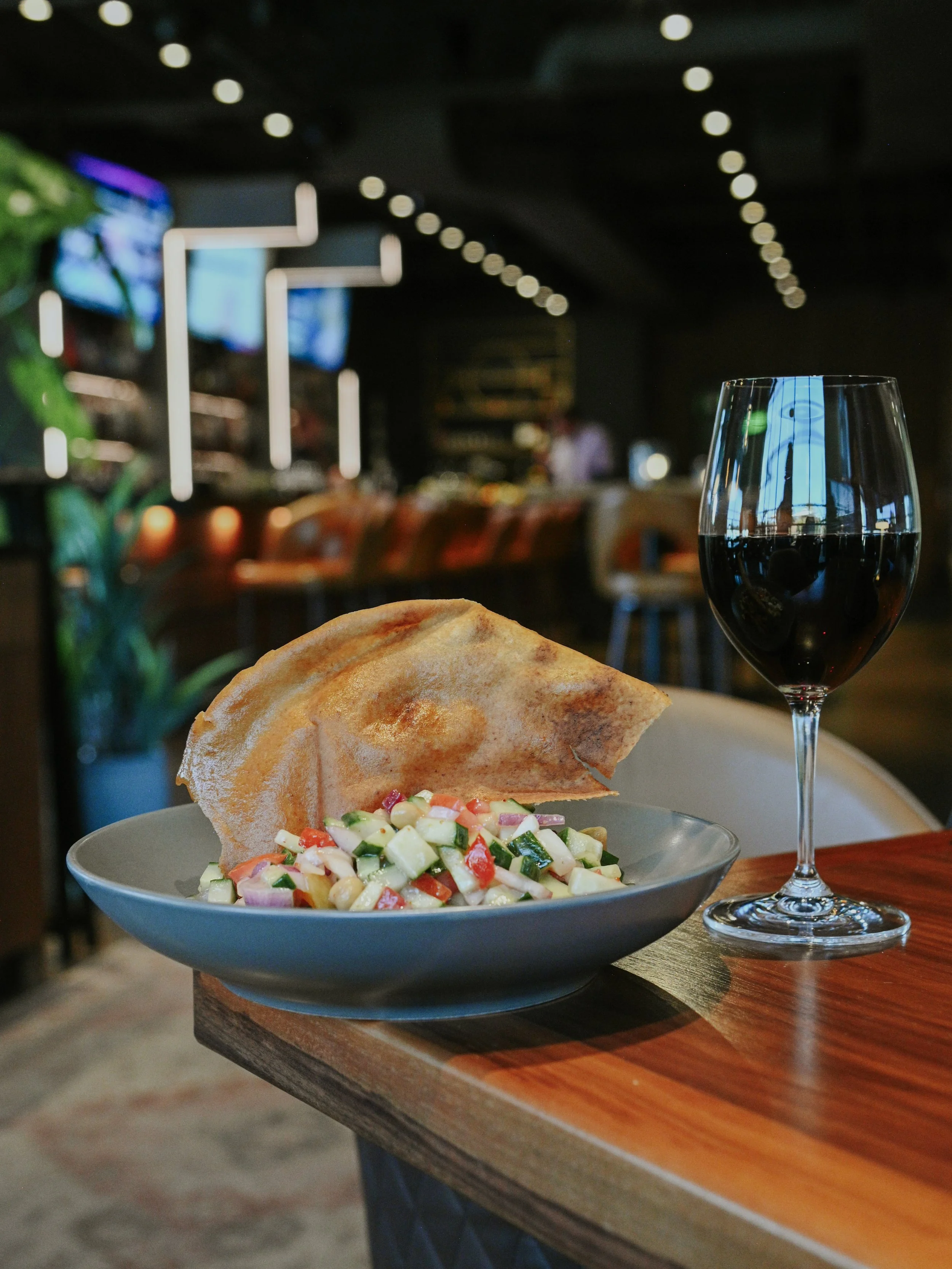 A plate of salad with diced vegetables and a piece of flatbread, accompanied by a glass of red wine on a wooden table in a restaurant.