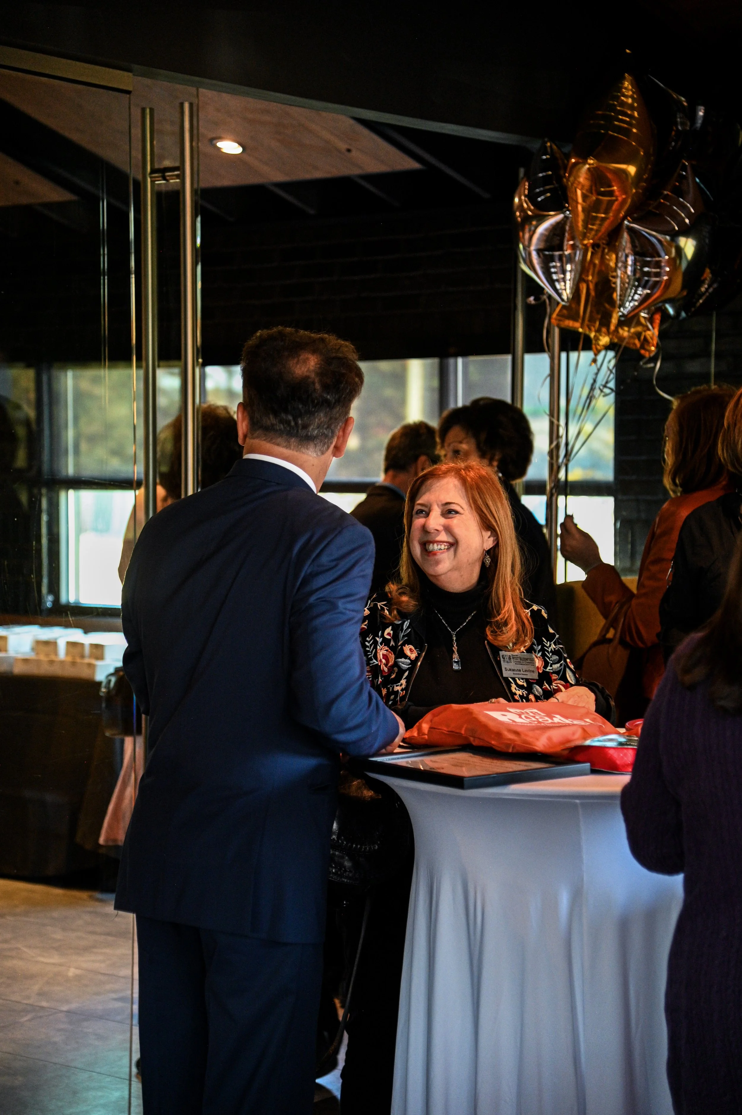 A woman with red hair, wearing a black top and floral jacket, smiling while talking to a man in a navy suit at a reception table decorated with balloons and a tote bag during a social event.