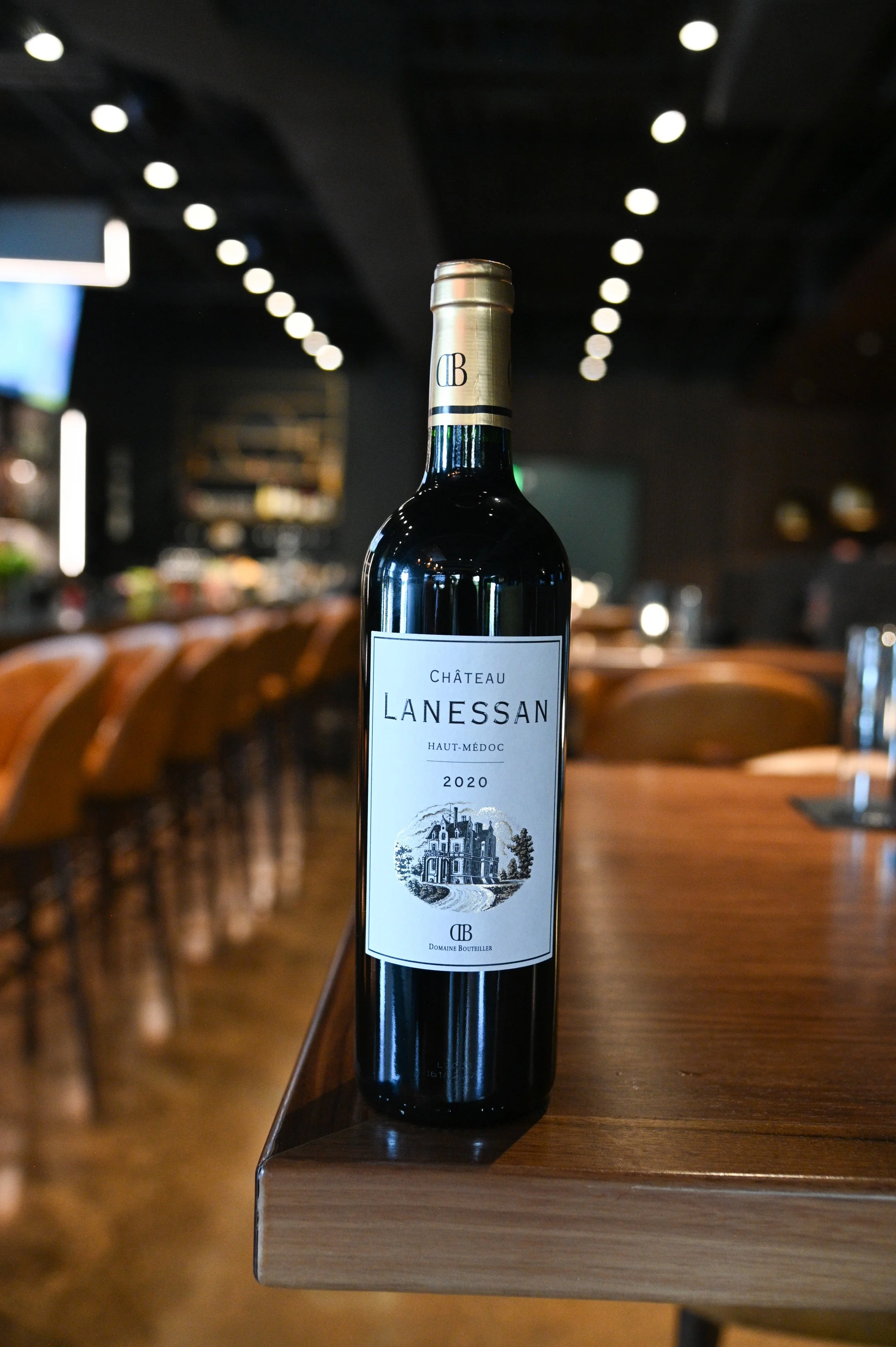 A bottle of Château Lanessan 2020 red wine is placed on a wooden table in a modern dining area with chairs and hanging lights.