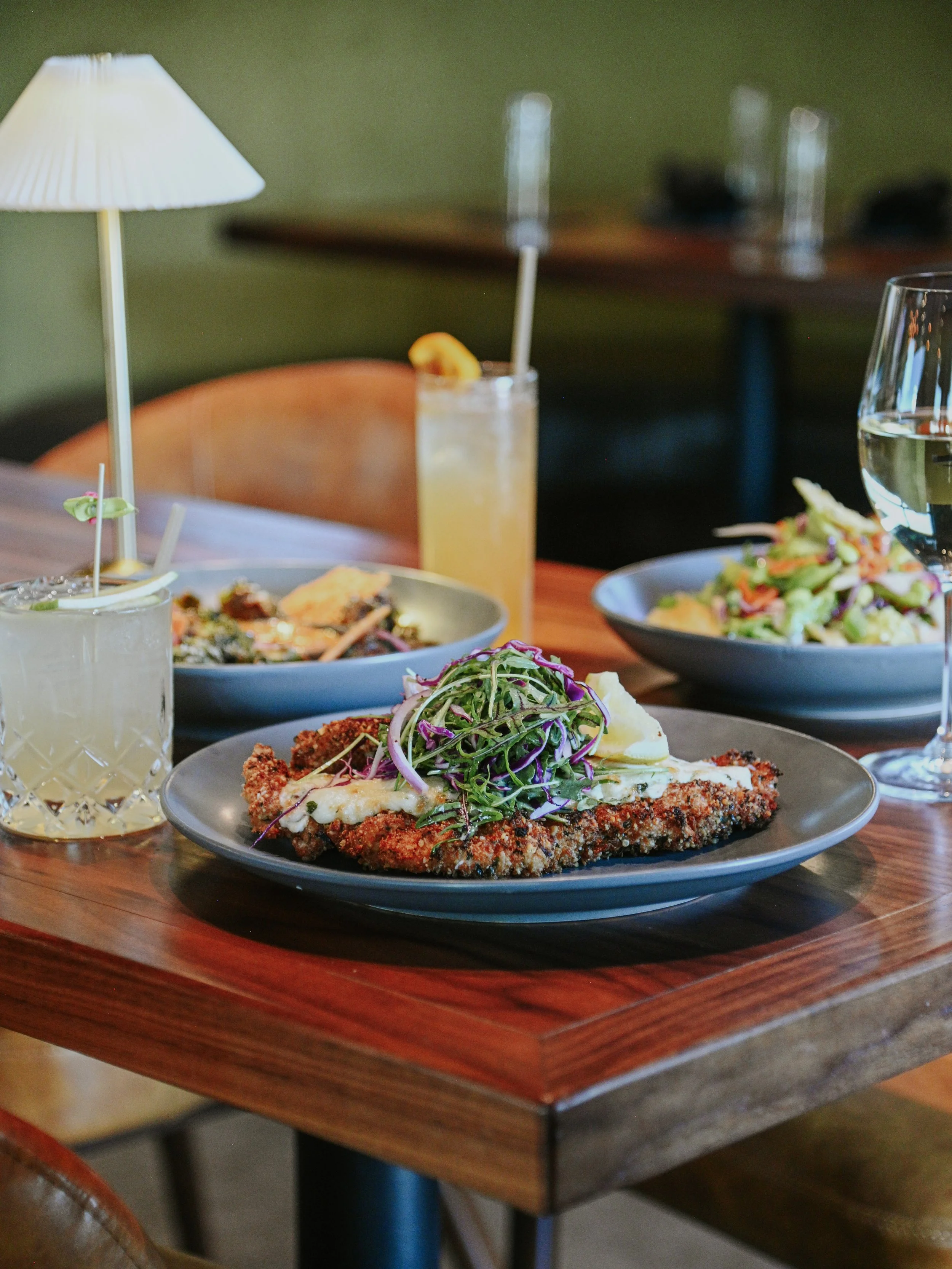 Plate with breaded meat topped with salad greens, on a wooden table with drinks and a salad in the background.