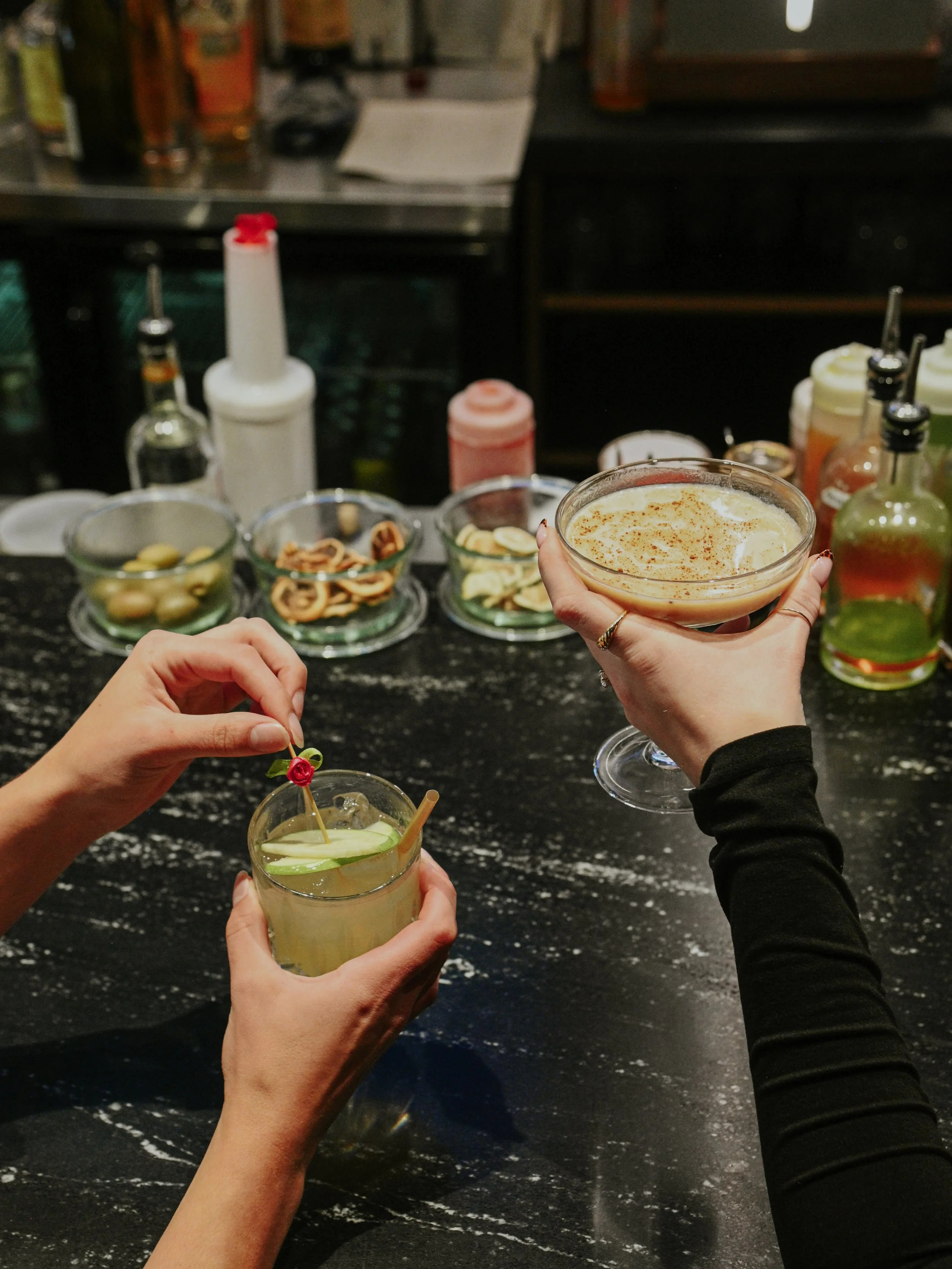 Two hands holding cocktails at a bar counter, with various bottles and snacks in the background.