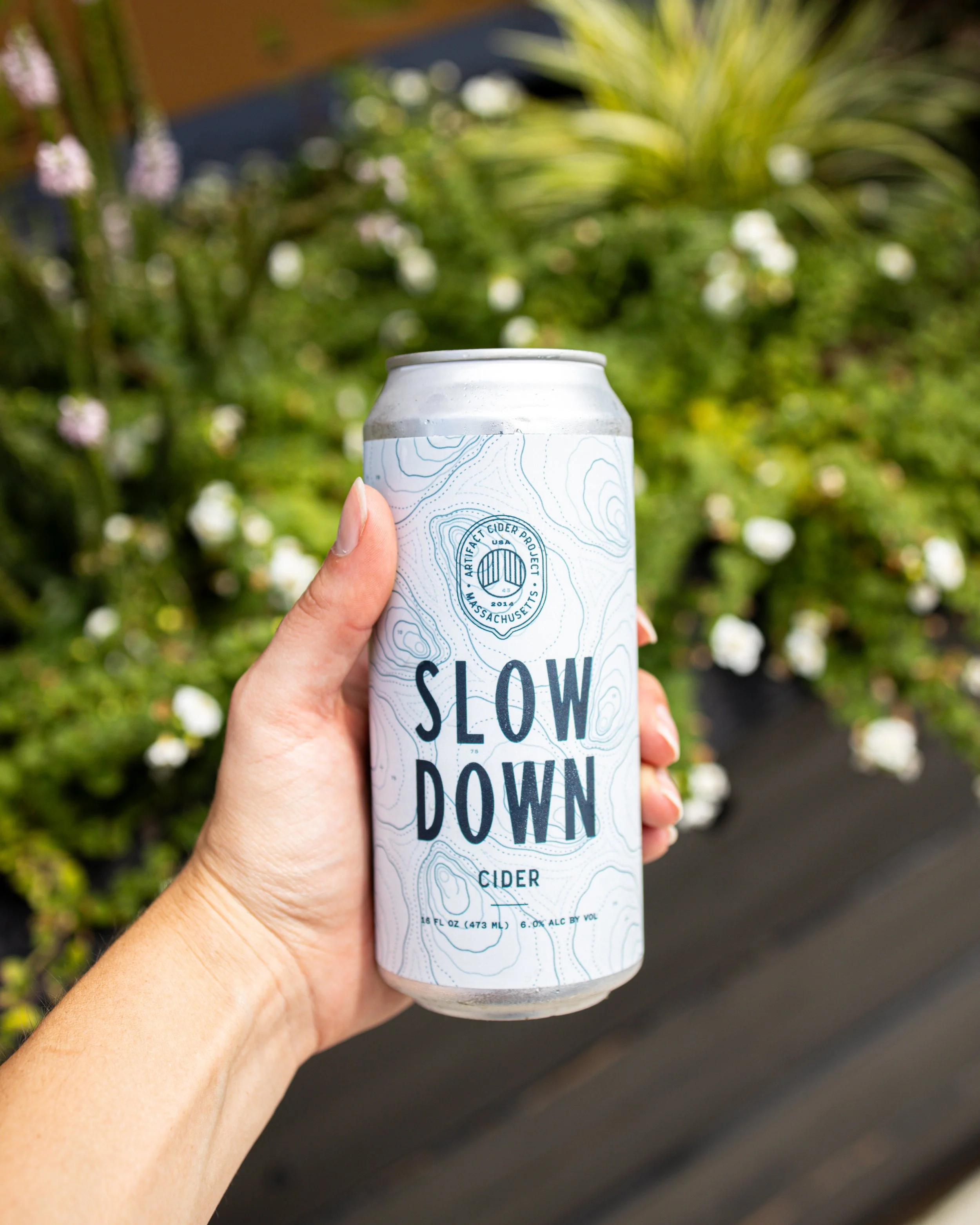 Hand holding a can of Slow Down cider in front of a garden with green plants and white flowers.