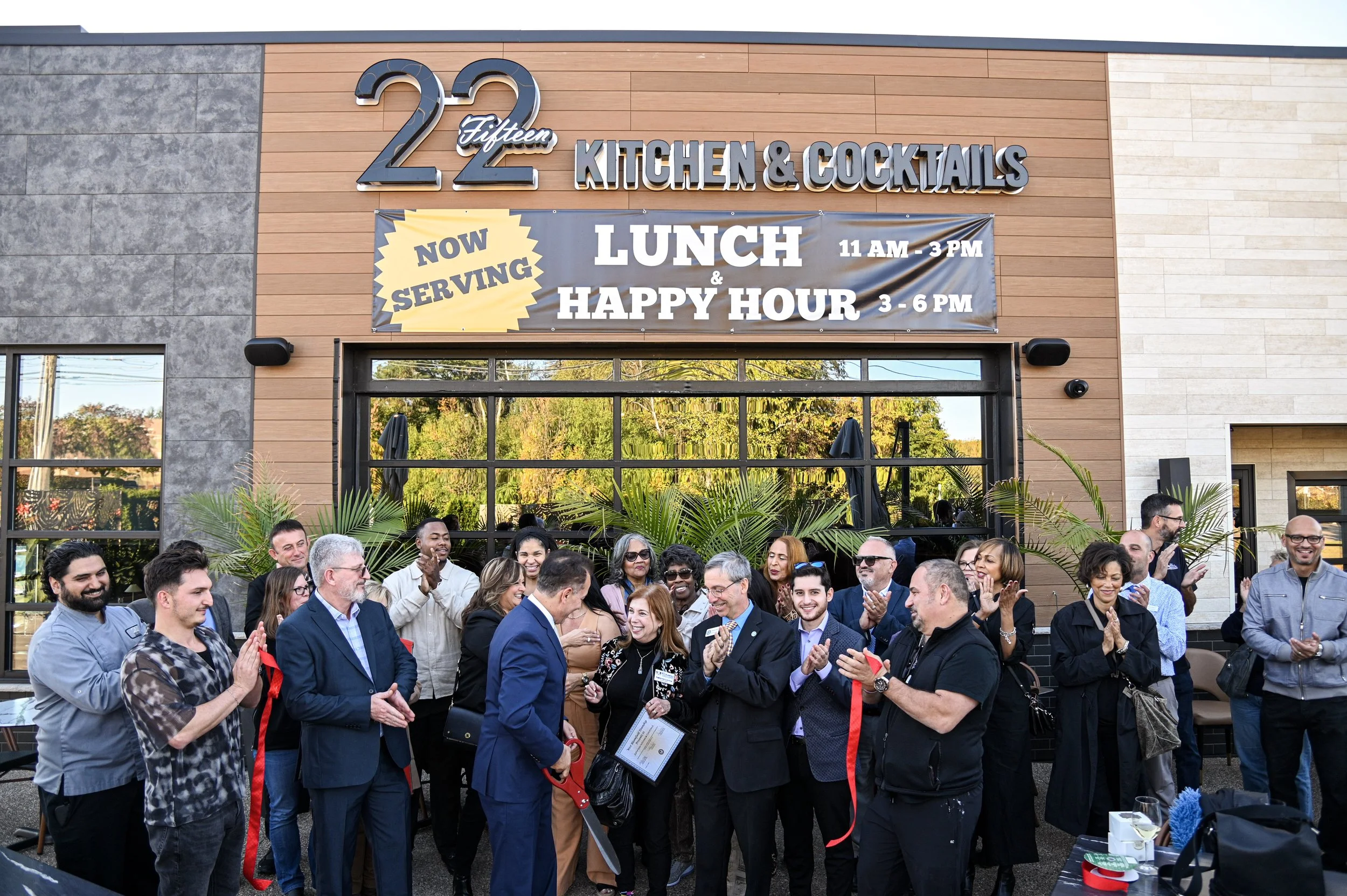 Group of people gathered outside a restaurant during a ribbon-cutting ceremony, with a sign advertising lunch and happy hour, in front of a building with a sign that reads '22 Fifteen Kitchen & Cocktails'.