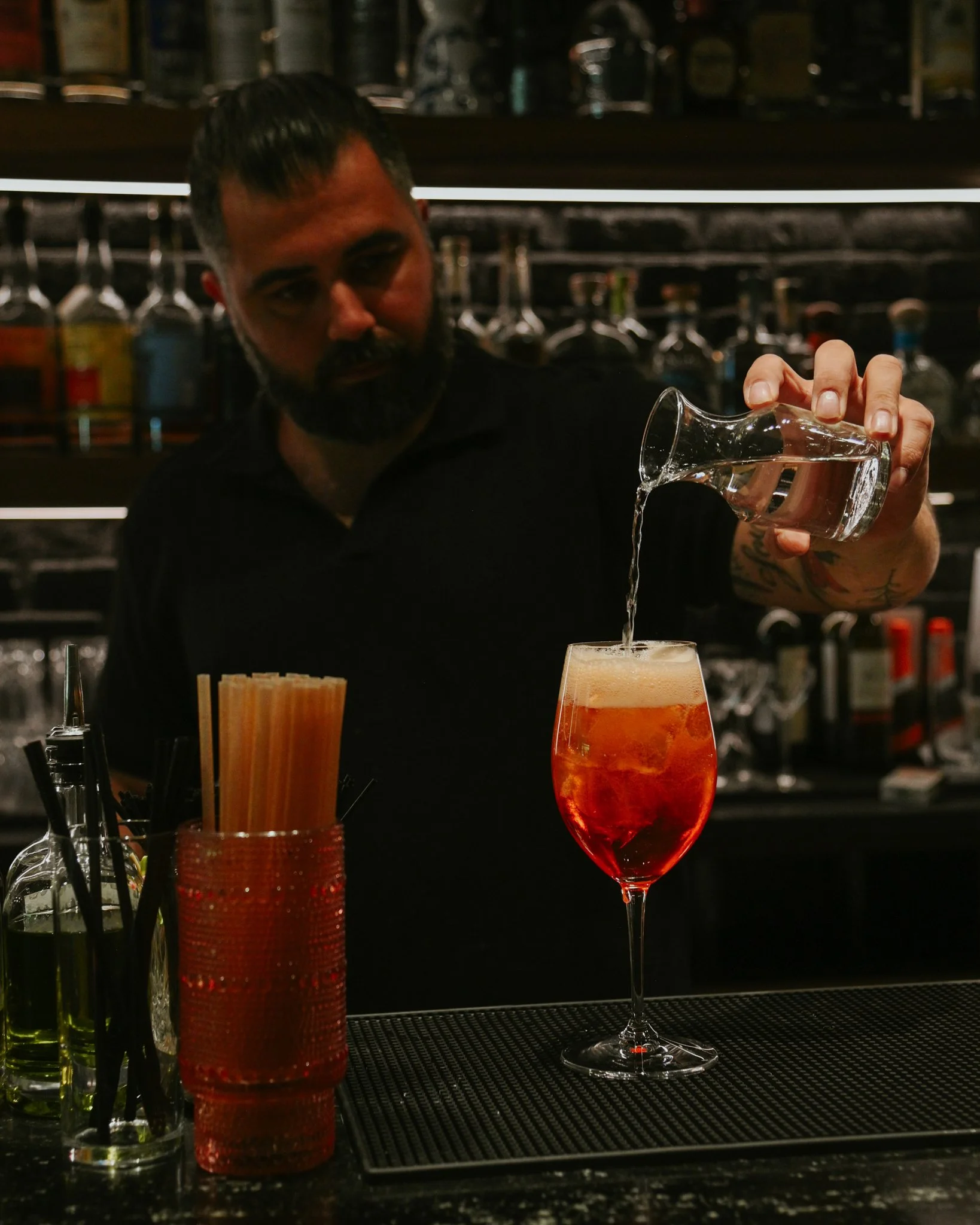 A bartender pouring water into a tall glass with a bright orange beverage on a black bar counter, with various bottles and bar tools in the background.