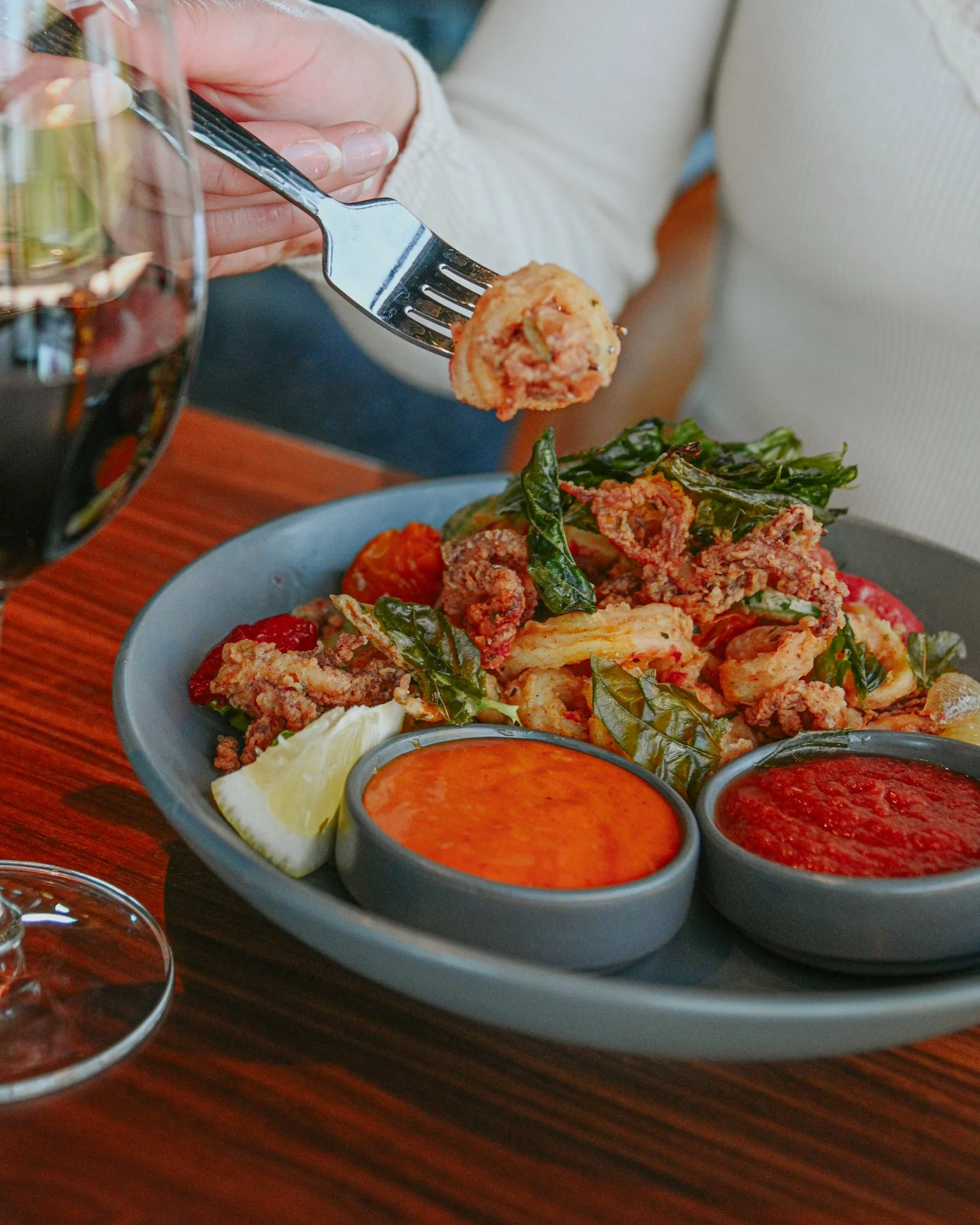 Person holding a fork with a piece of fried food over a plate of fried squid, shiso leaves, cherry tomatoes, lemon wedge, and two small bowls of red dipping sauces on a wooden table.