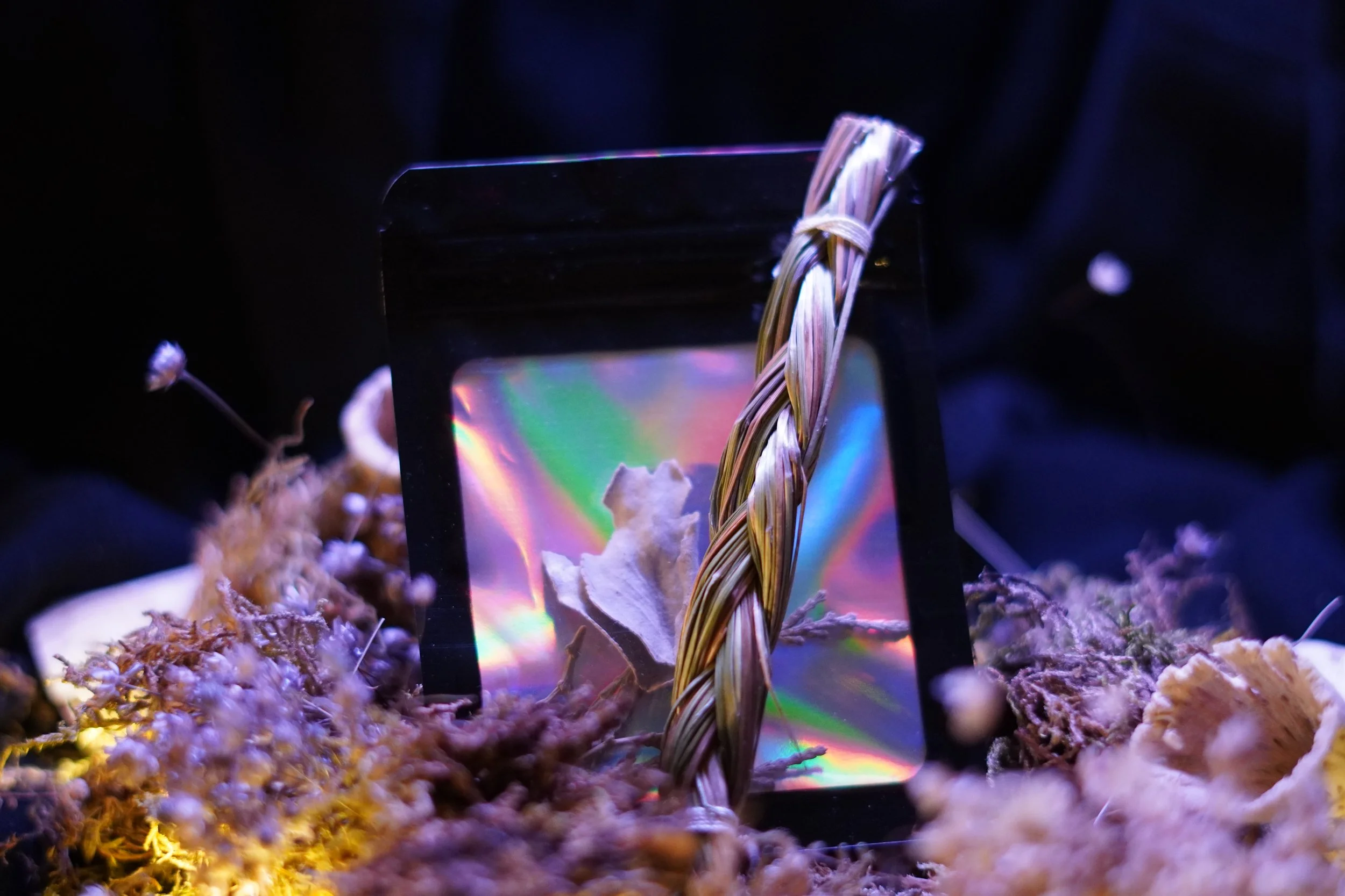 A braid of sweetgrass pictured next to sage and cedar inside a multicolored sealed packet, on top of moss and dried flowers 