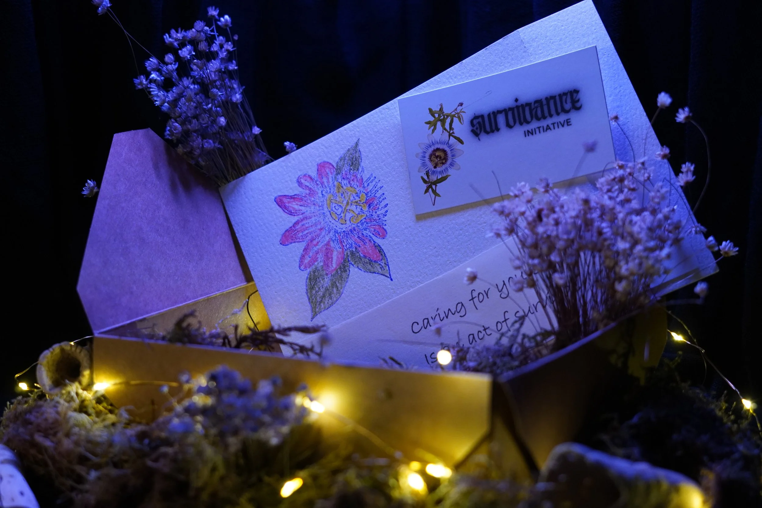 A handmade card and sticker sit within a cardstock box, amongst dried flowers, moss, and tiny lights
