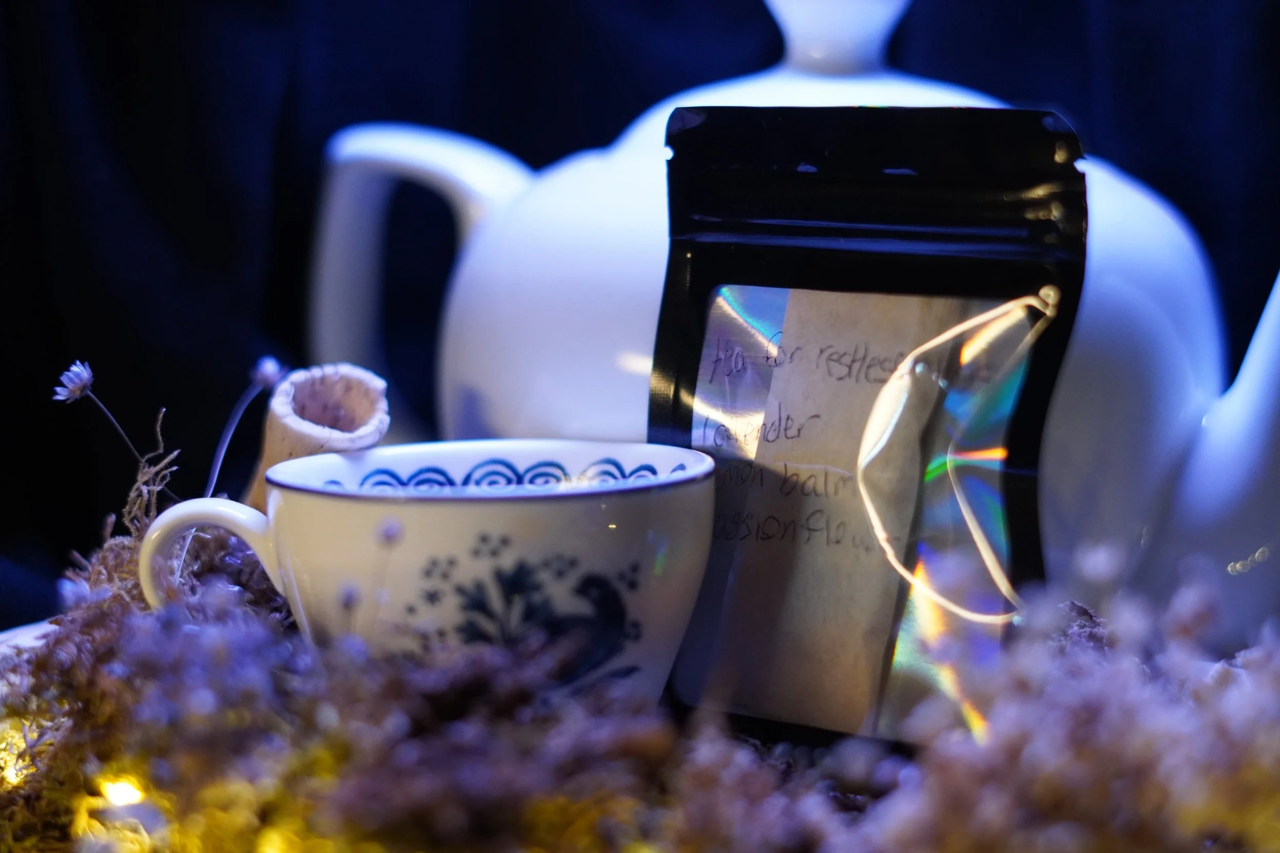A sealed packed marked tea for restlessness leans on a white tea pot and cop, sitting on dried moss and flowers