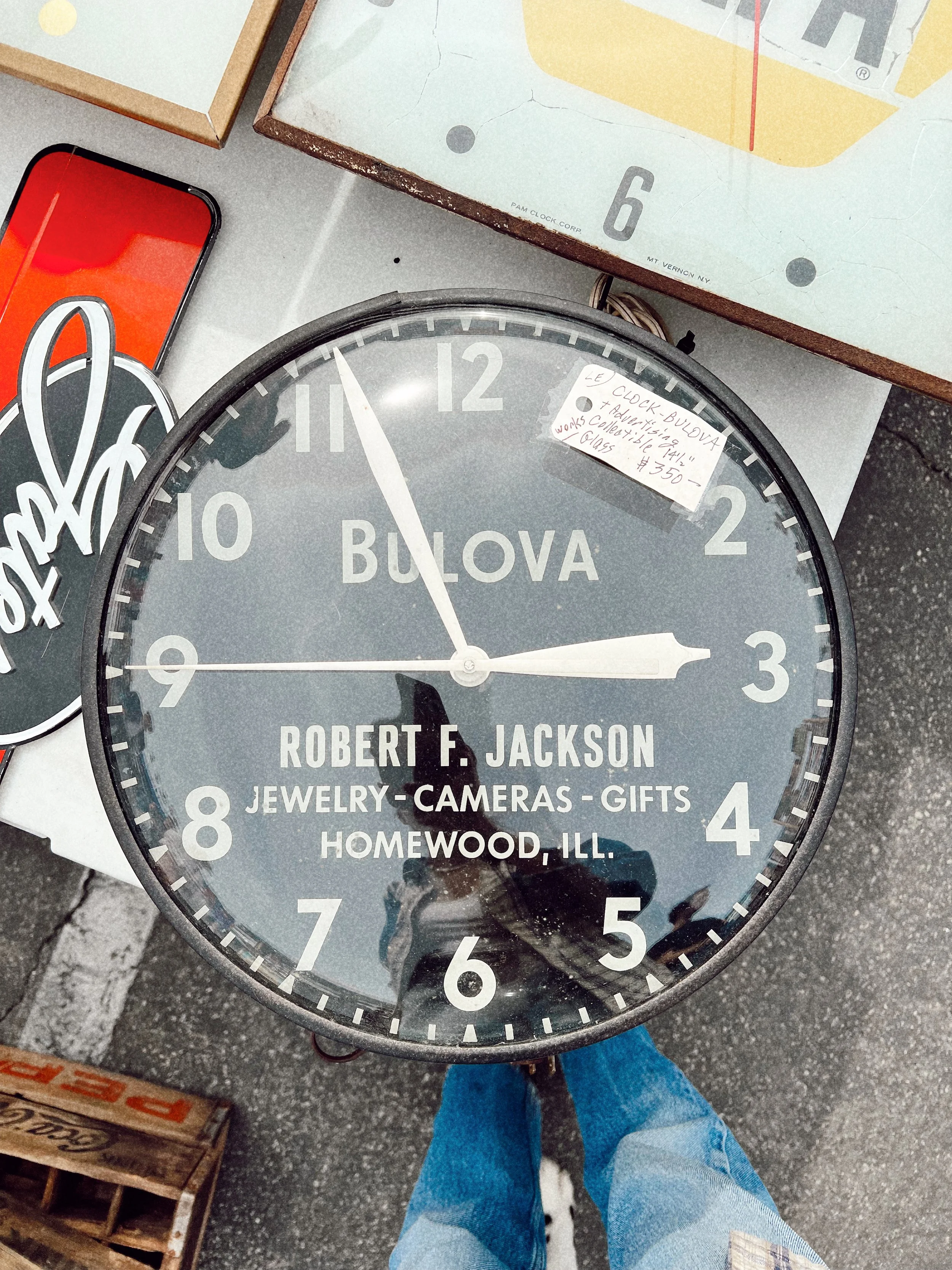 A large vintage Bulova wall clock showing 2:58, placed on a surface among various other items including a smartphone, signs, and boxes. The clock face has text indicating it belongs to Robert F. Jackson, involved in jewelry, cameras, and gifts, locat