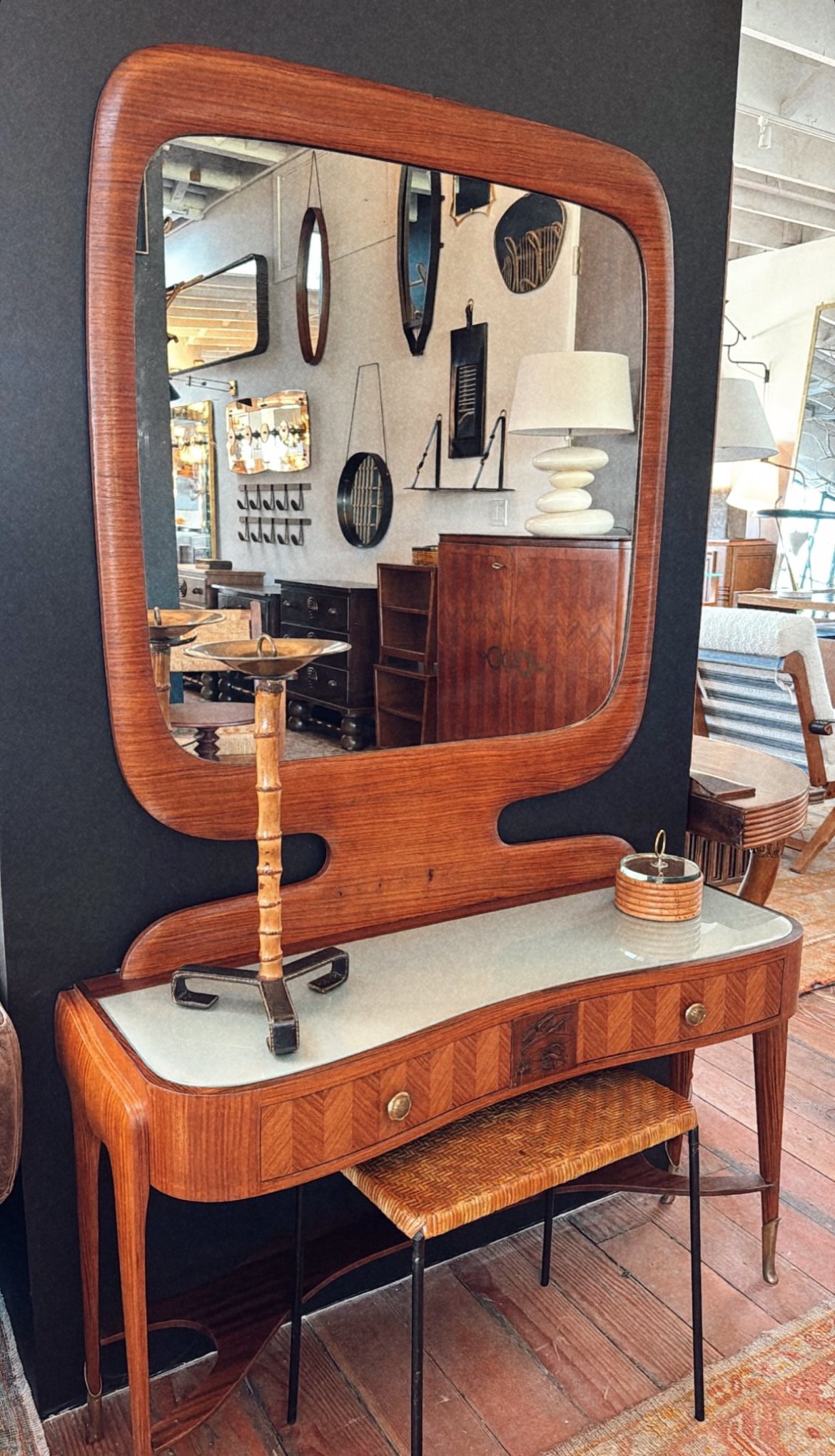A vintage wooden dressing table with a large mirror, a small sculptural table lamp, and a cane stool in a furniture store. Reflection shows various wall decor items.