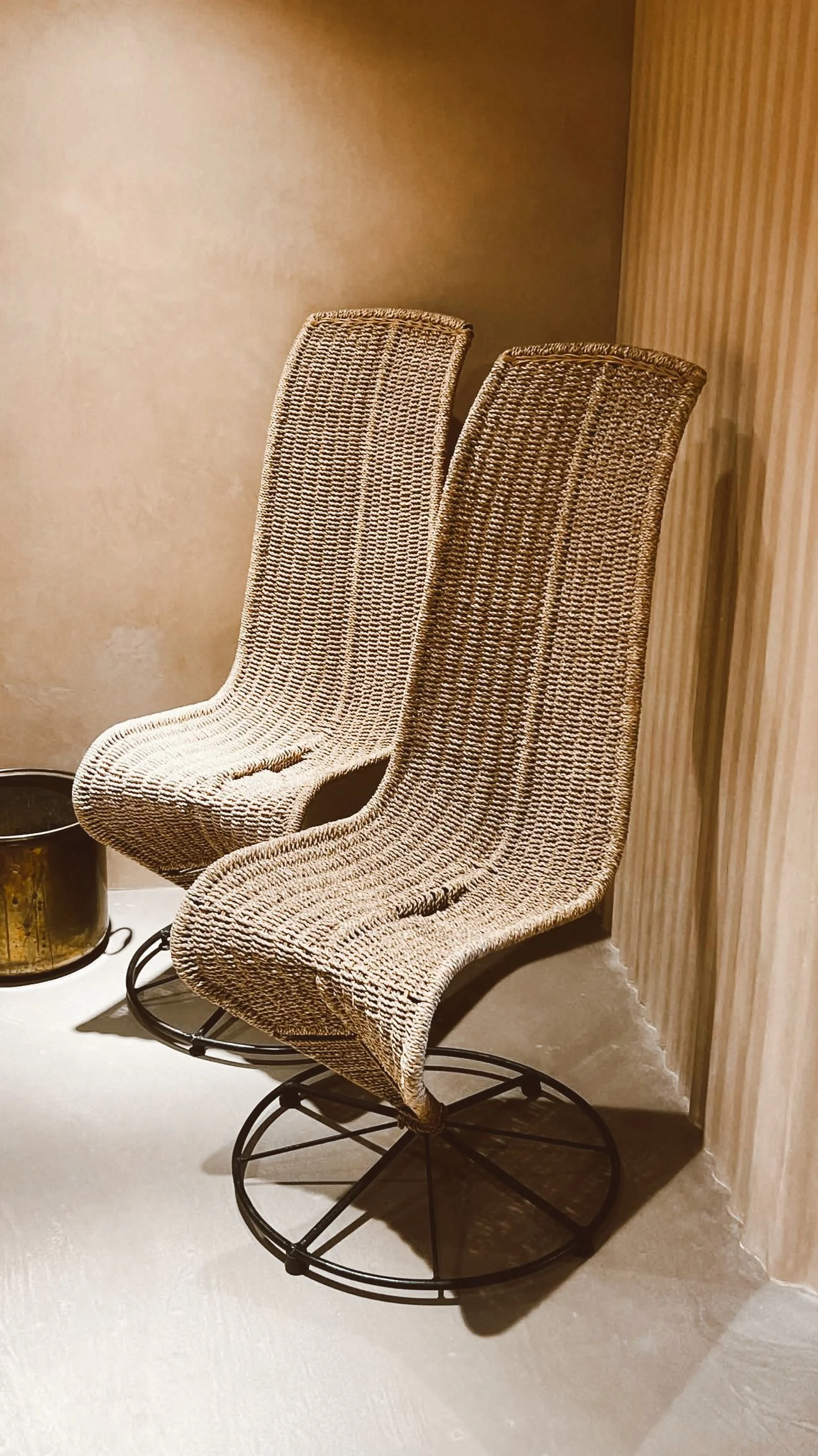 Two woven rattan bar stools with metal circular bases, placed in a corner beside a textured wall.