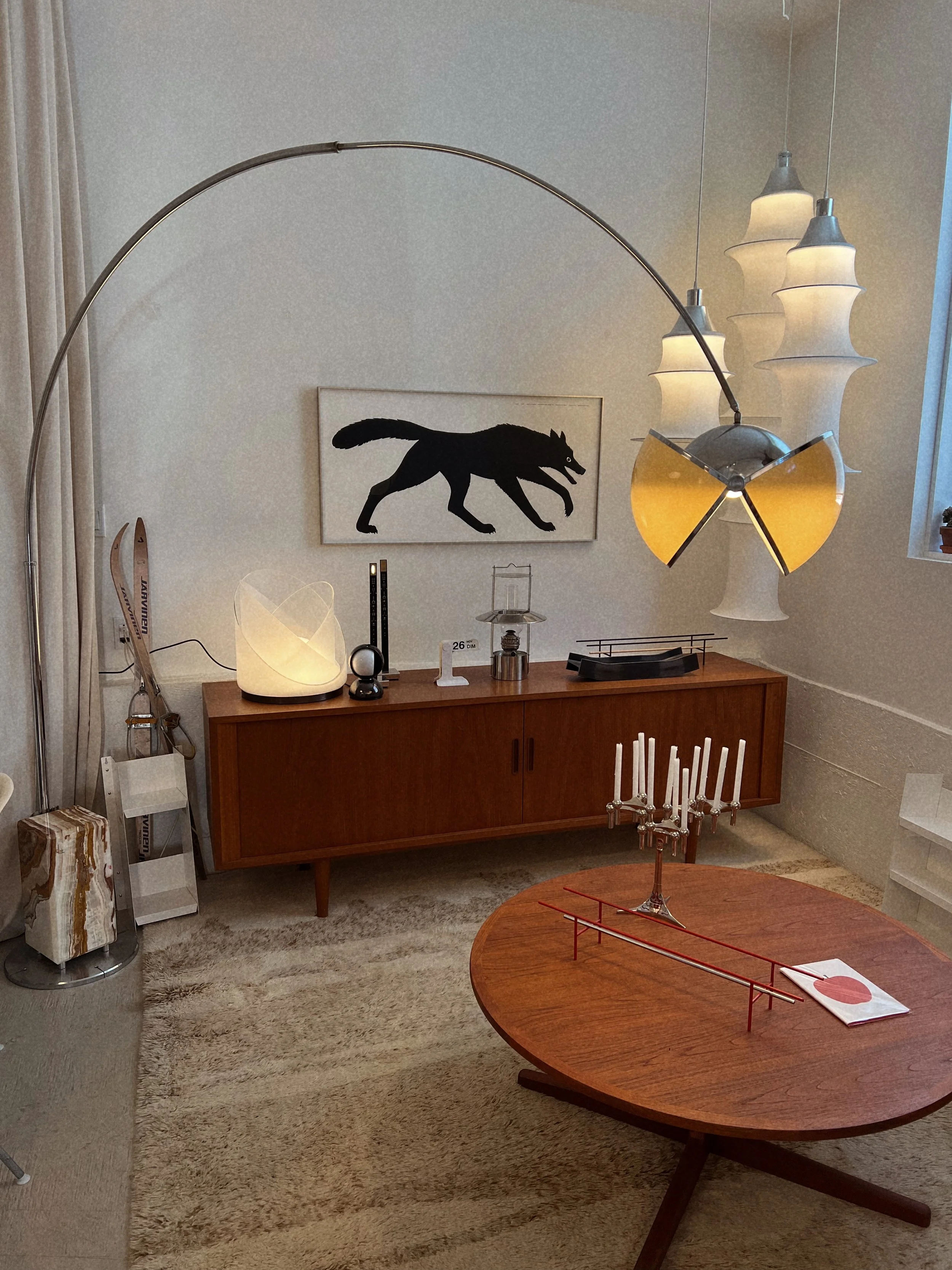 A mid-century modern living room with a wooden sideboard, a circular wooden table, a framed black silhouette of a wolf, a white sculptural lamp, hanging pendant lights, stock skis, and a candelabra on the table.