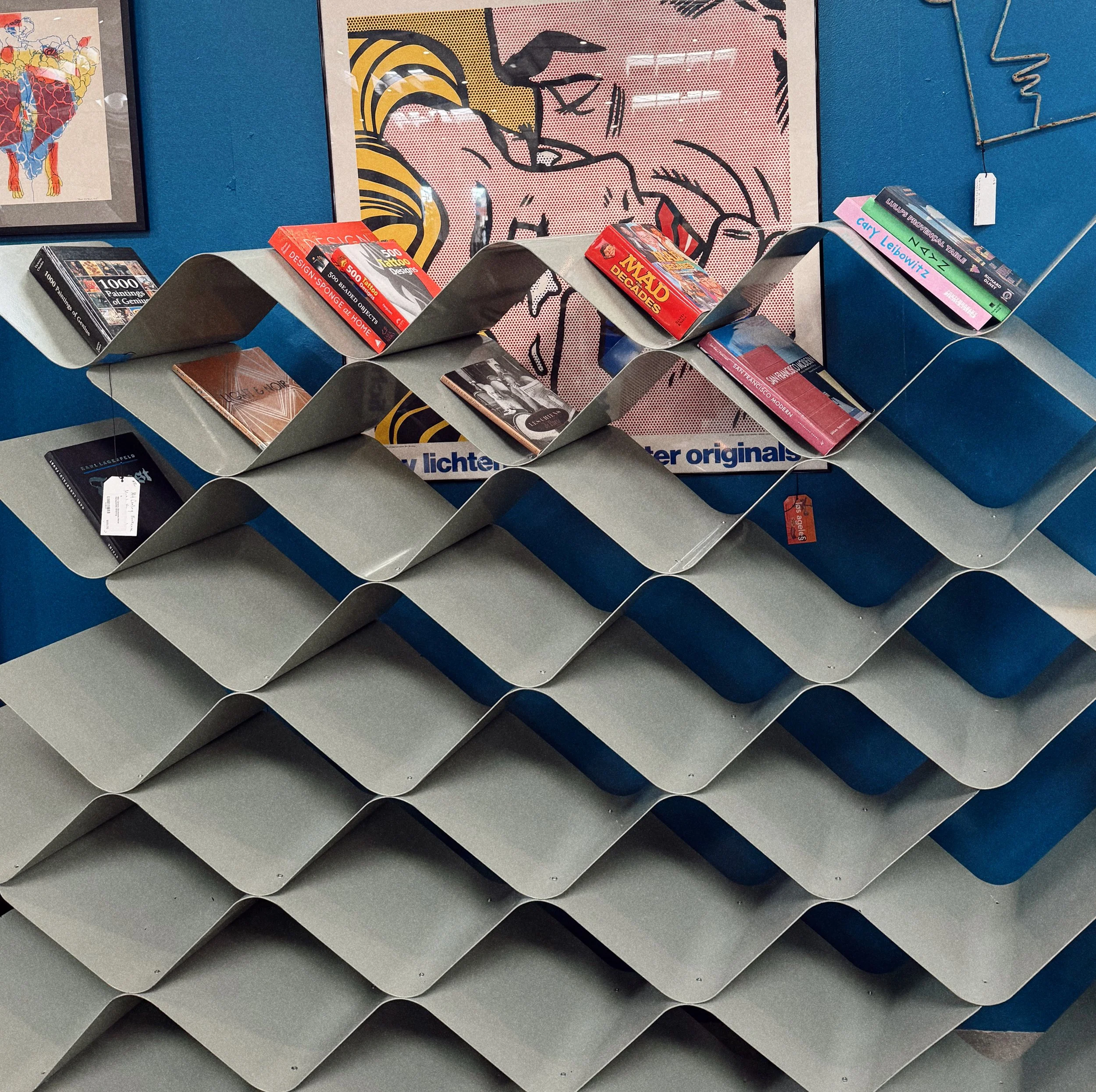 Empty gray diagonal wall-mounted shelves with magazines and books on top, against a blue wall with artwork.