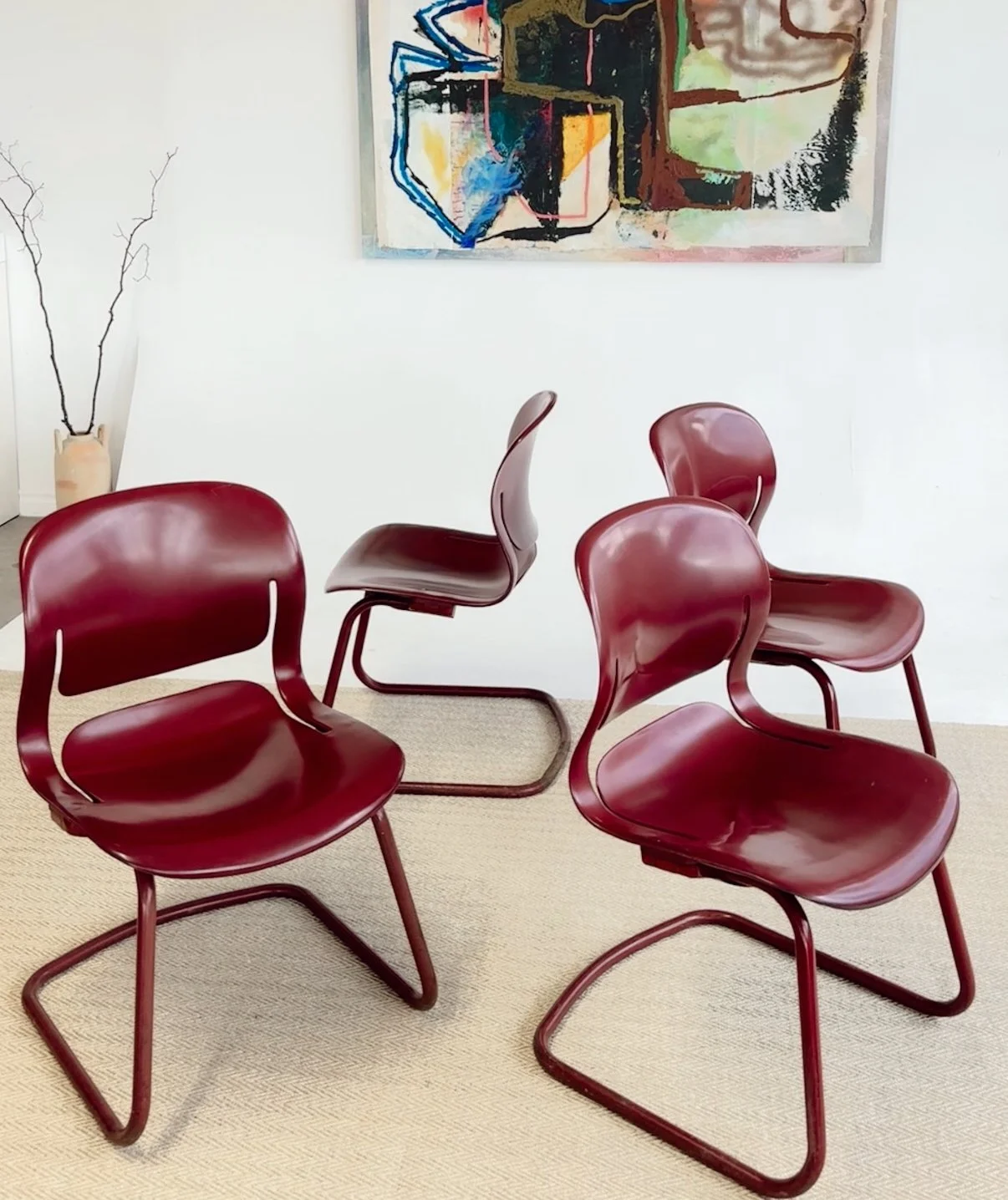 Four red mid-century modern chairs with metal frames arranged in a staggered pattern in front of a white wall with abstract artwork and a vase with decorative branches.