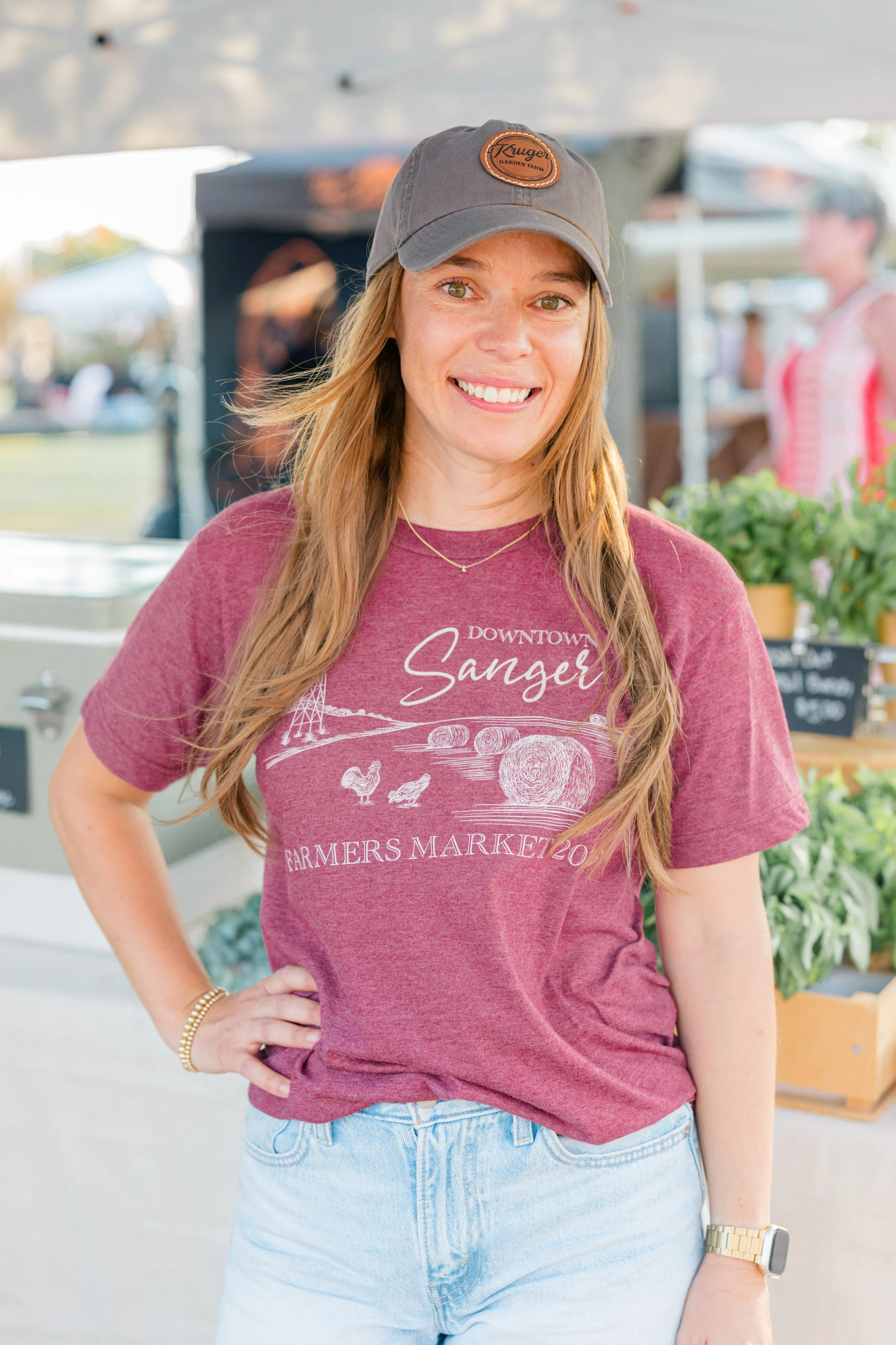 Young woman smiling at a farmers' market stand with jars of honey, fresh strawberries in small brown containers, and a chalkboard sign displaying prices, under a white canopy.