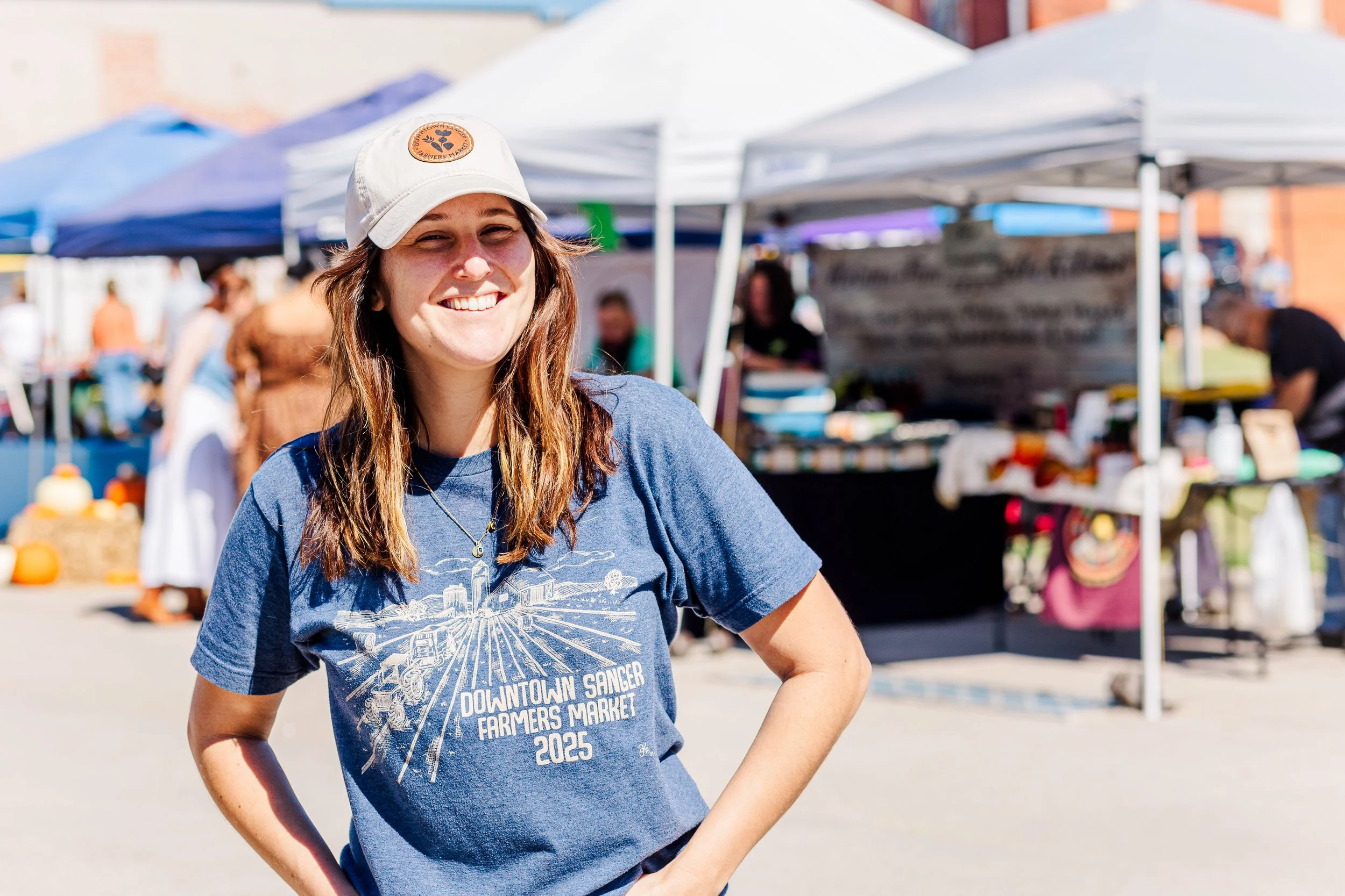 Two people sitting at a table at an outdoor farmers market, smiling, with tote bags reading 'Valley View Farmers Market' in front of them, in a park setting with a gazebo in the background.